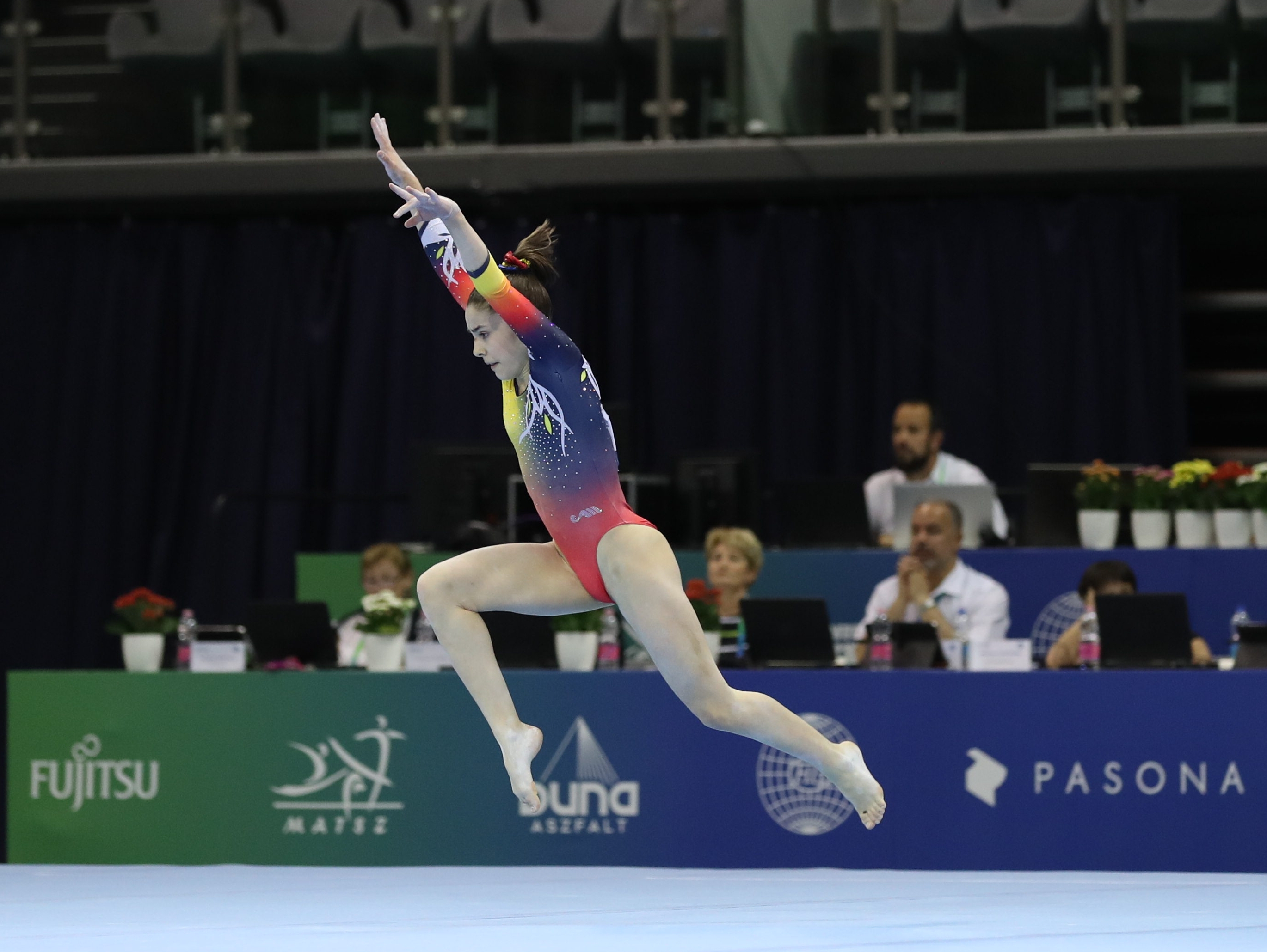 A gymnast leaps high in the air during a floor exercise routine at an indoor competition.