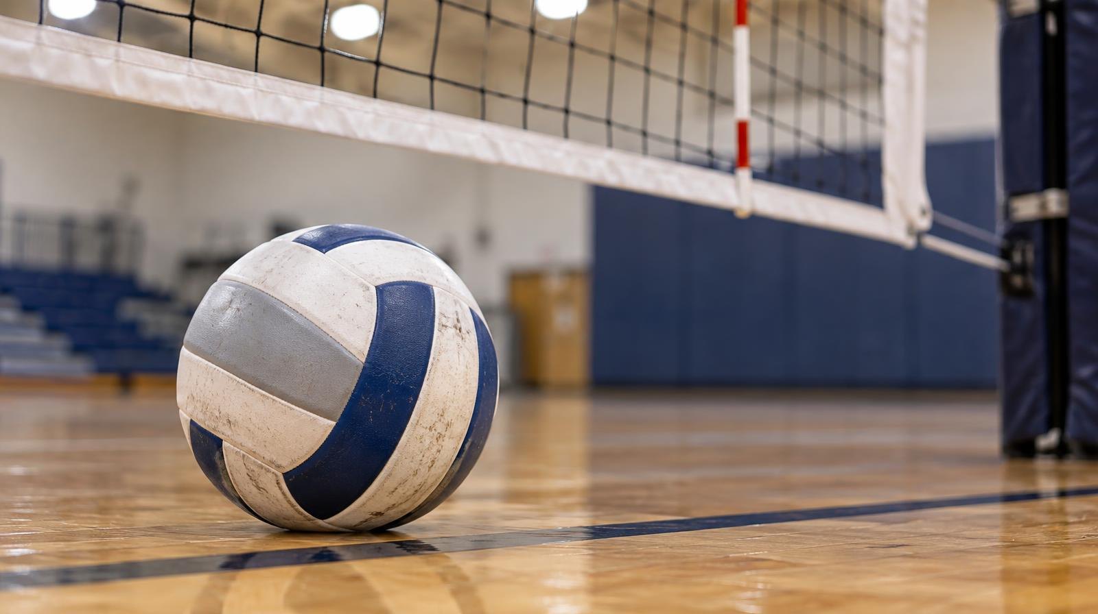 A volleyball resting on a hardwood gym floor near a volleyball net.