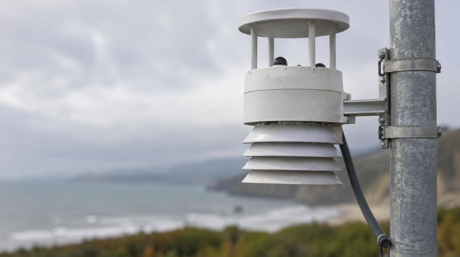 Outdoor air quality monitoring sensor on a metal pole against a hazy sky.