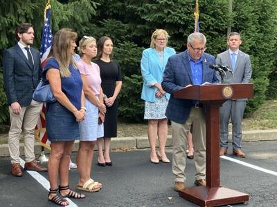 A man speaks at an outdoor podium bearing an official seal, flanked by a group of people near an American flag.