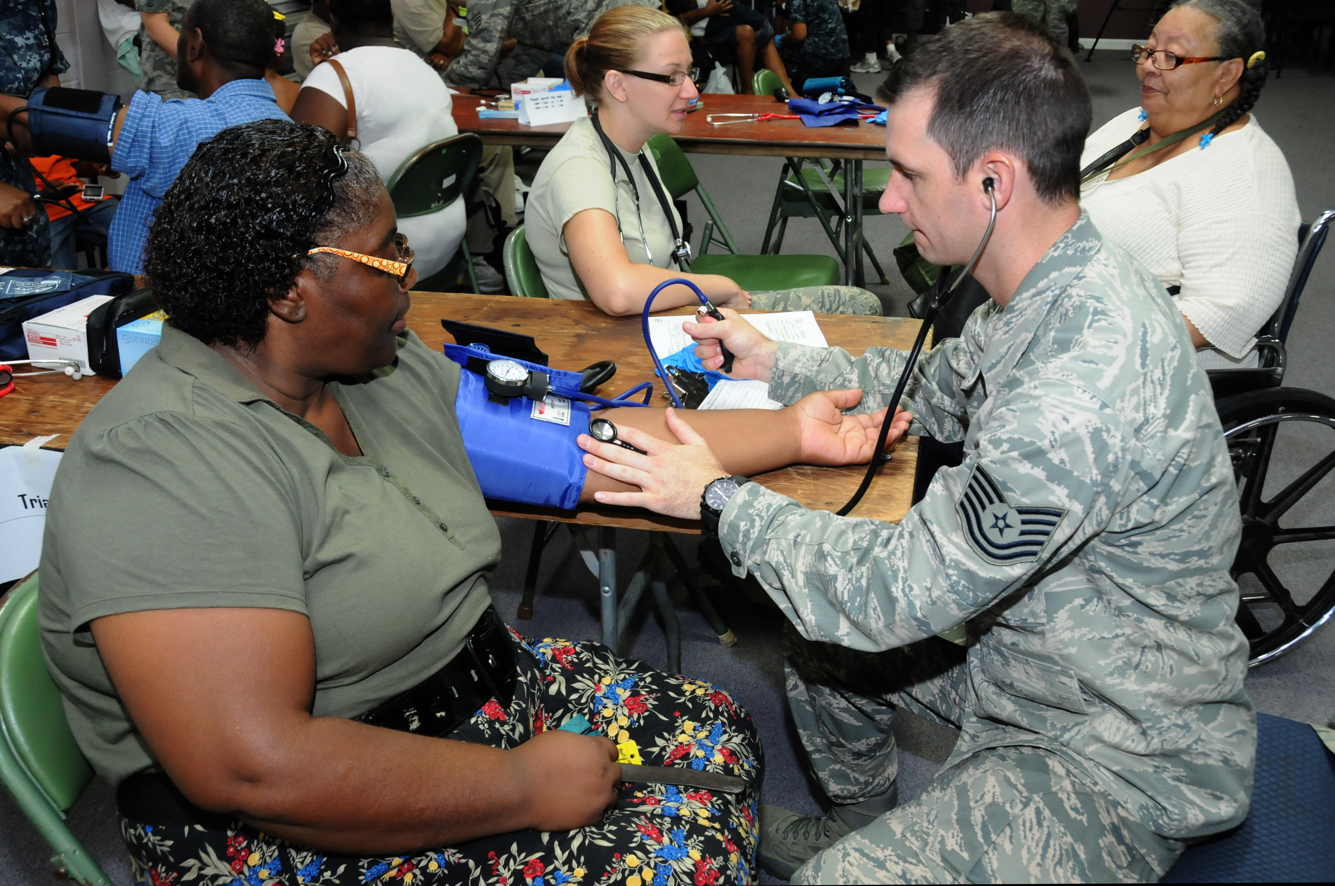 A uniformed service member checks a civilian woman's blood pressure at a community health screening.