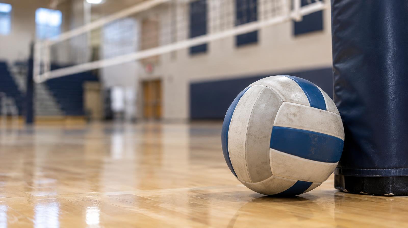 A volleyball resting on a hardwood gym floor next to a net post.