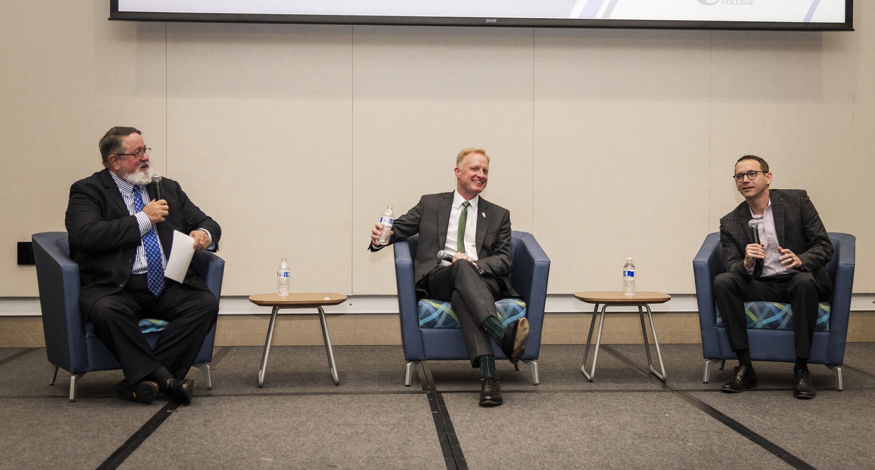 Three men in business attire seated in a row at a panel discussion event.