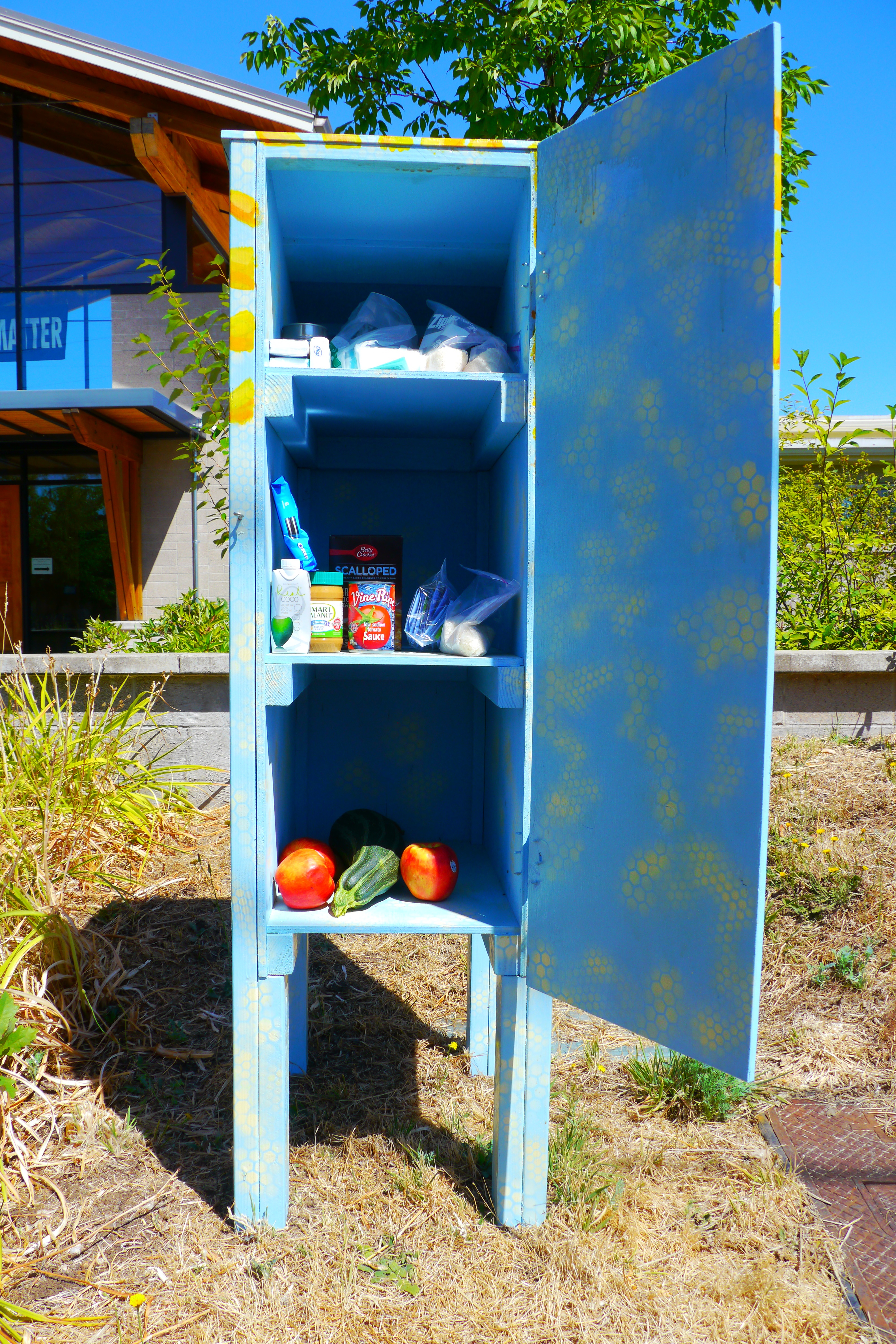 Open blue outdoor food pantry cabinet stocked with canned goods and fresh produce.
