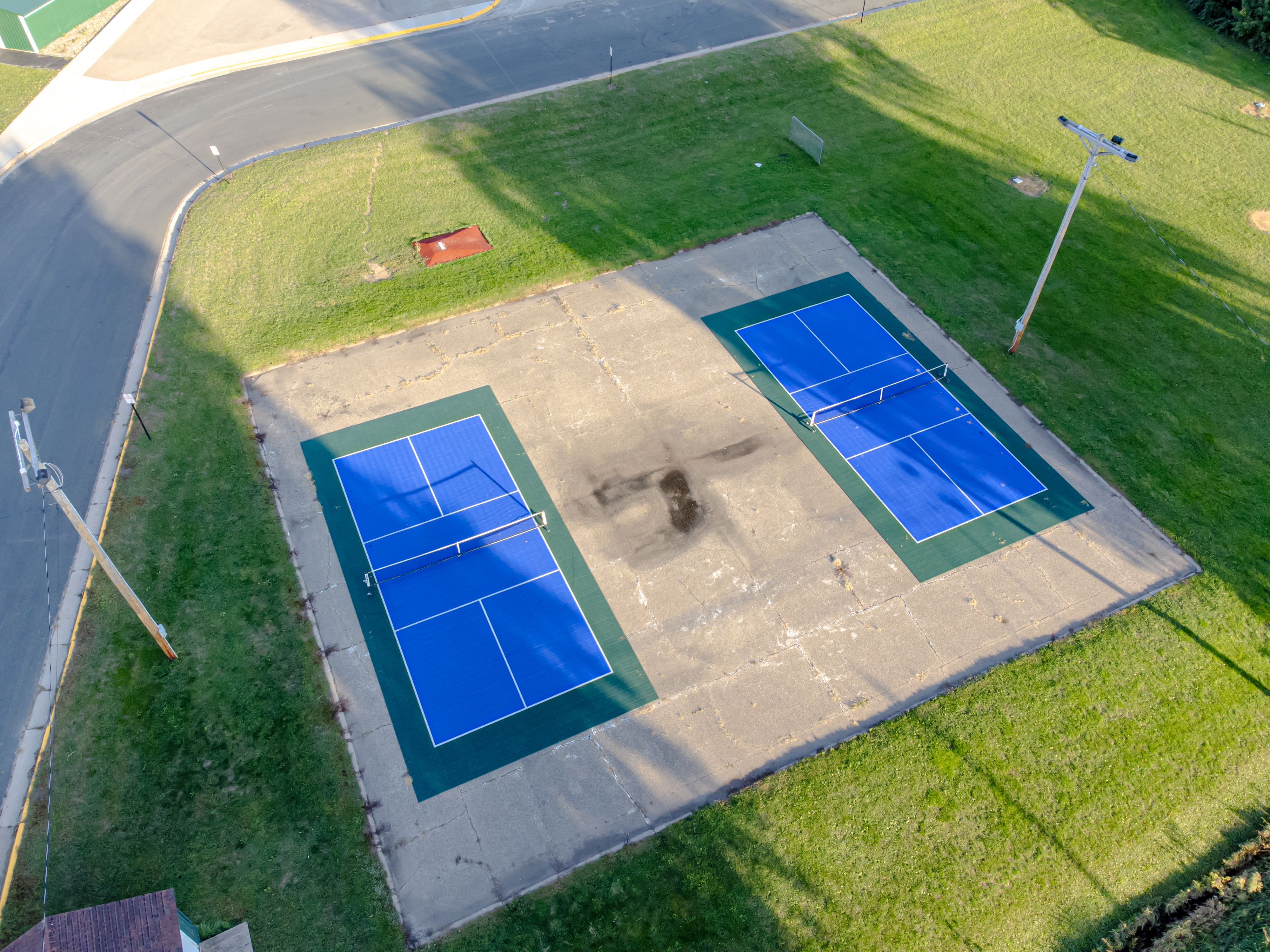 Aerial view of two blue outdoor pickleball courts with nets, surrounded by green grass and a curved road.