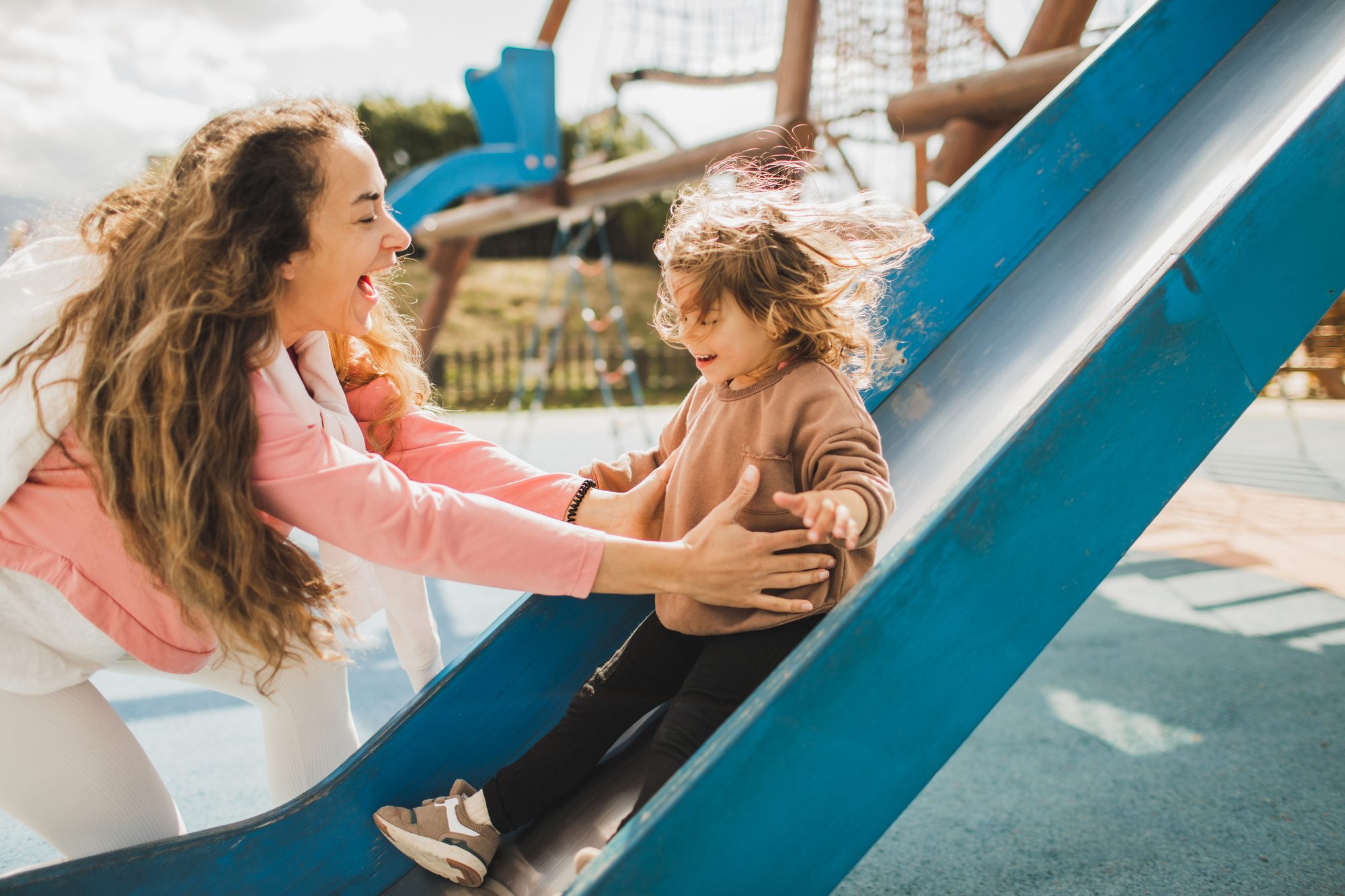 Laughing mom catching her happy daughter sliding on kids playground