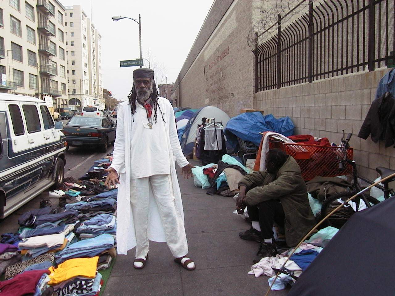 Tents, tarps, and belongings spread along an urban sidewalk near parked cars.