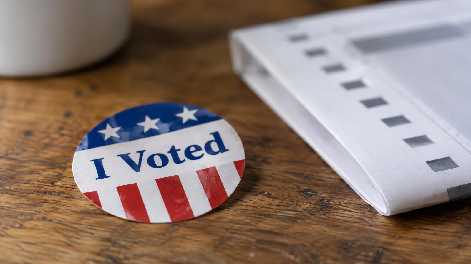 A red, white, and blue I Voted sticker resting on a wooden surface beside a folded ballot.