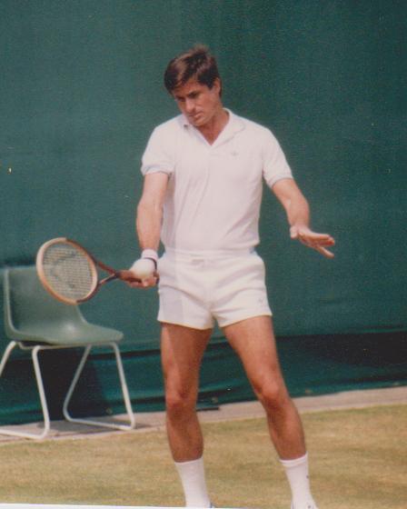 Man in white tennis attire swinging a wooden racket on a grass court