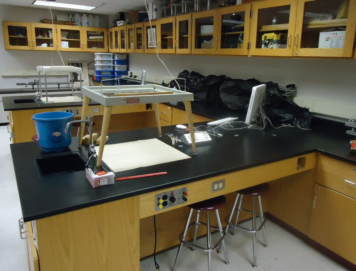 High school science classroom with black lab benches, stools, and wooden cabinetry.
