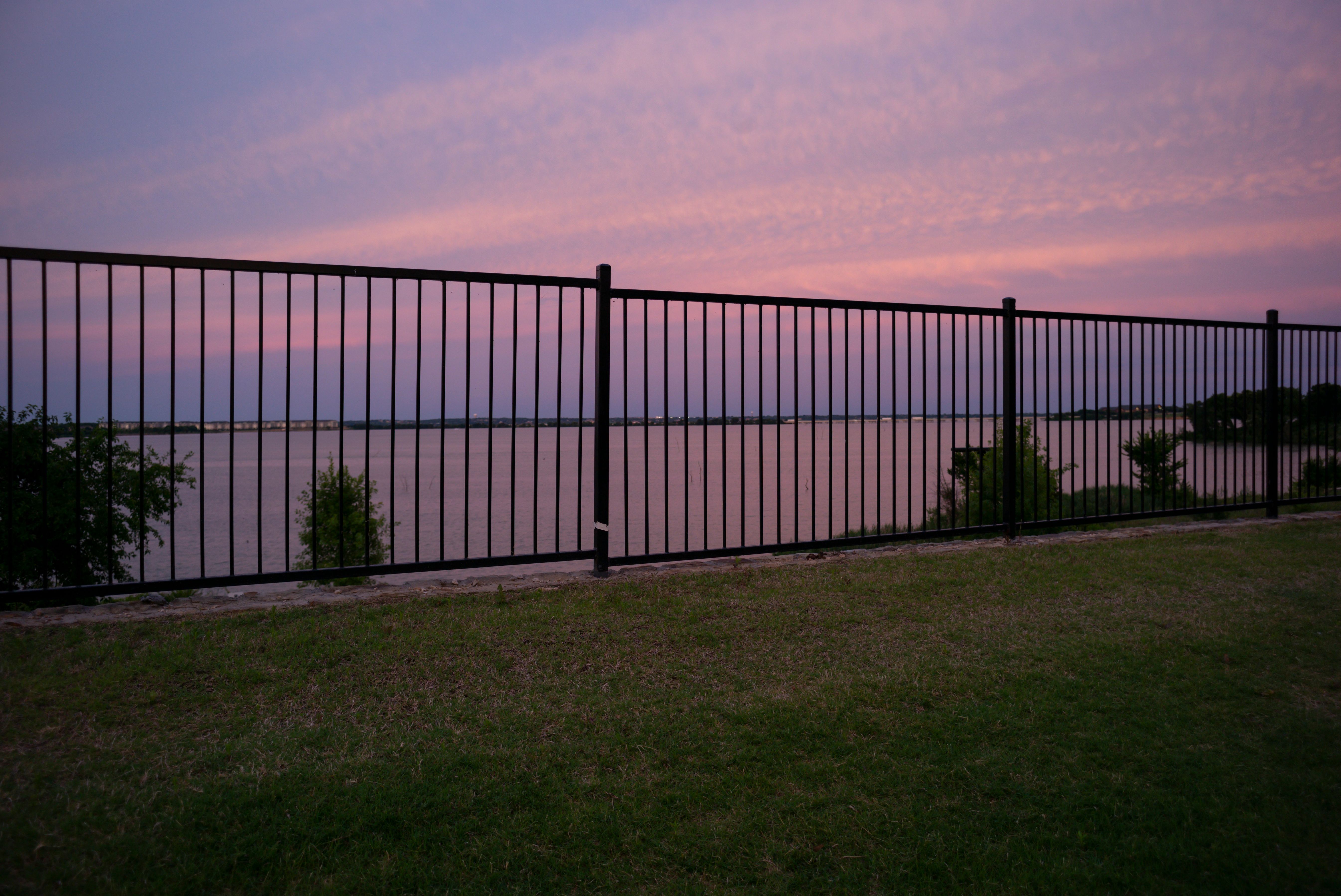 Black iron fence along a grassy shore overlooking a wide lake at sunset.