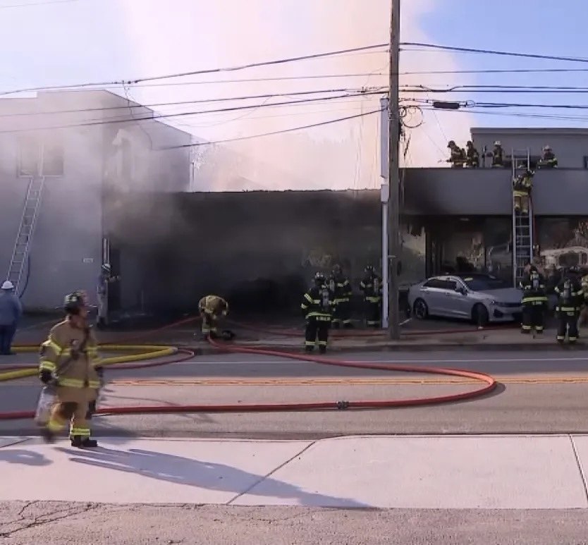 Firefighters in gear battle heavy smoke at a burning commercial building with hose lines deployed.