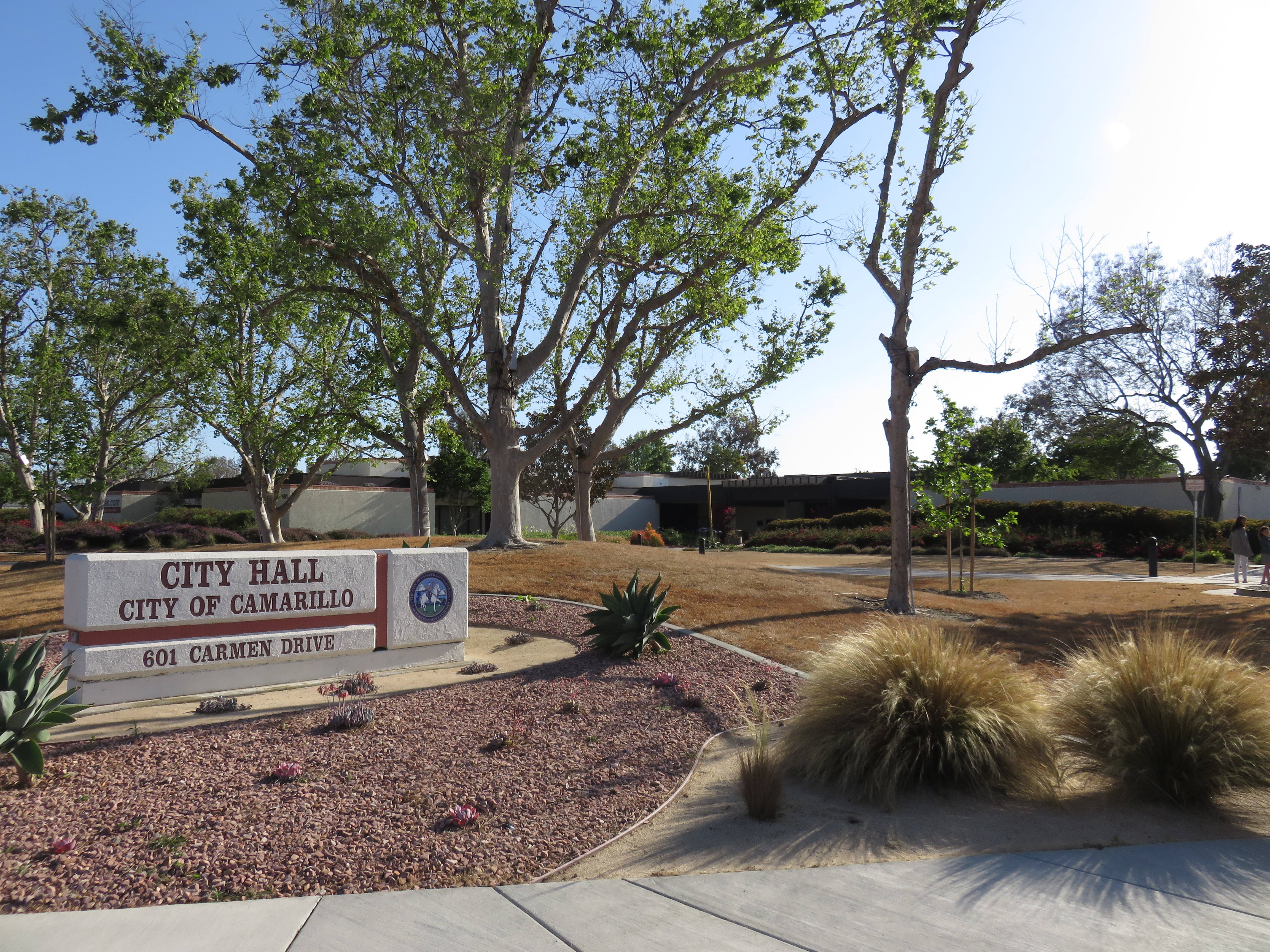 Sign and exterior of a low municipal building surrounded by trees and landscaping.