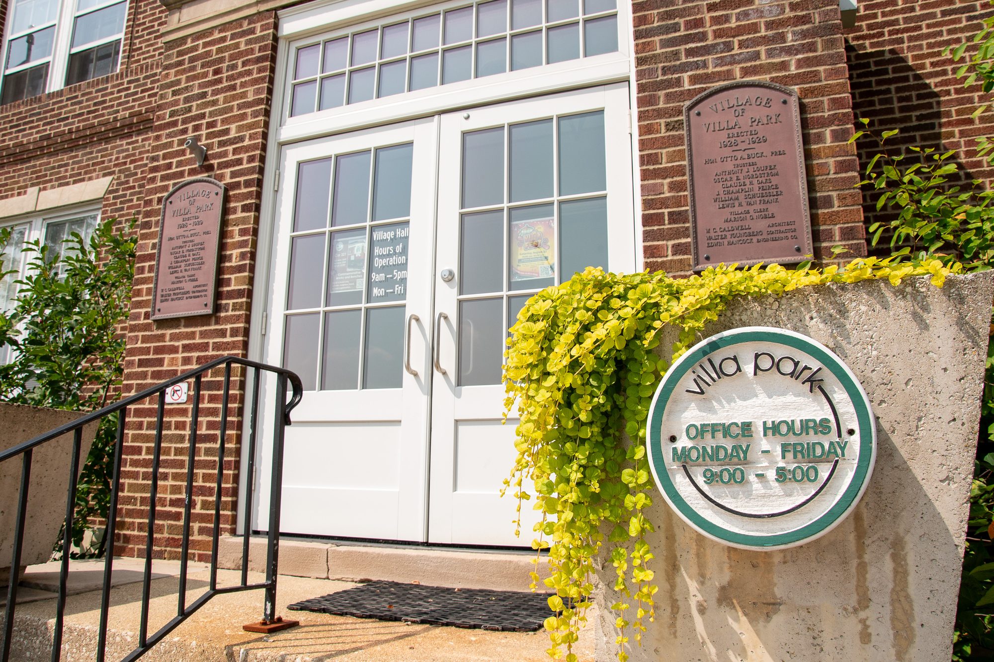 Brick municipal building entrance with white double doors and a stone village sign.