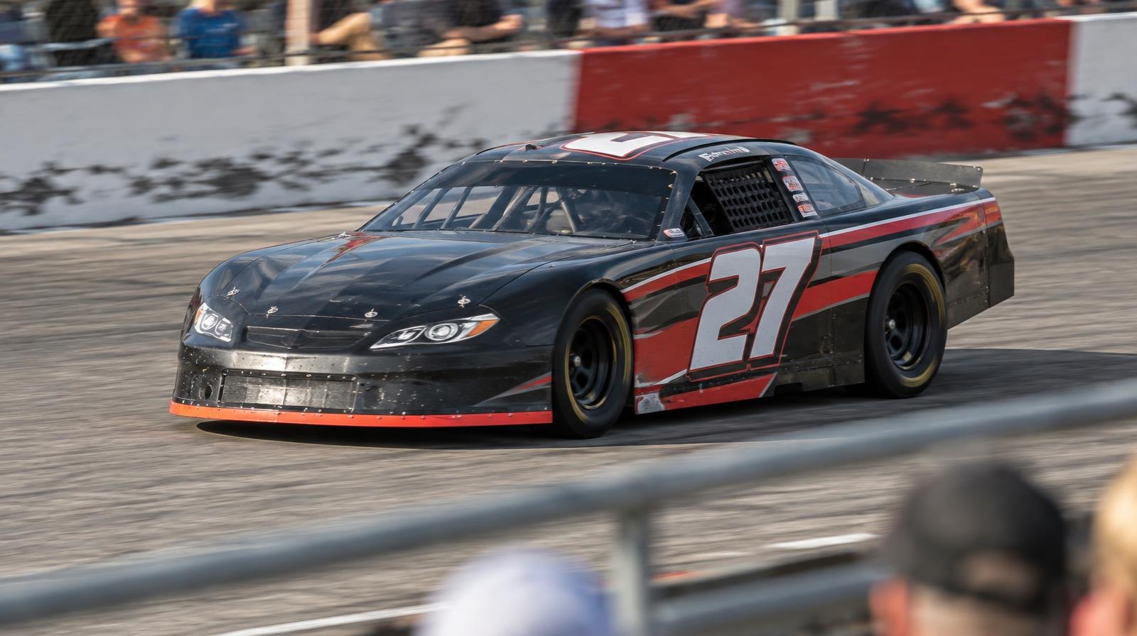 A colorful stock car races on a short oval track on a sunny afternoon.