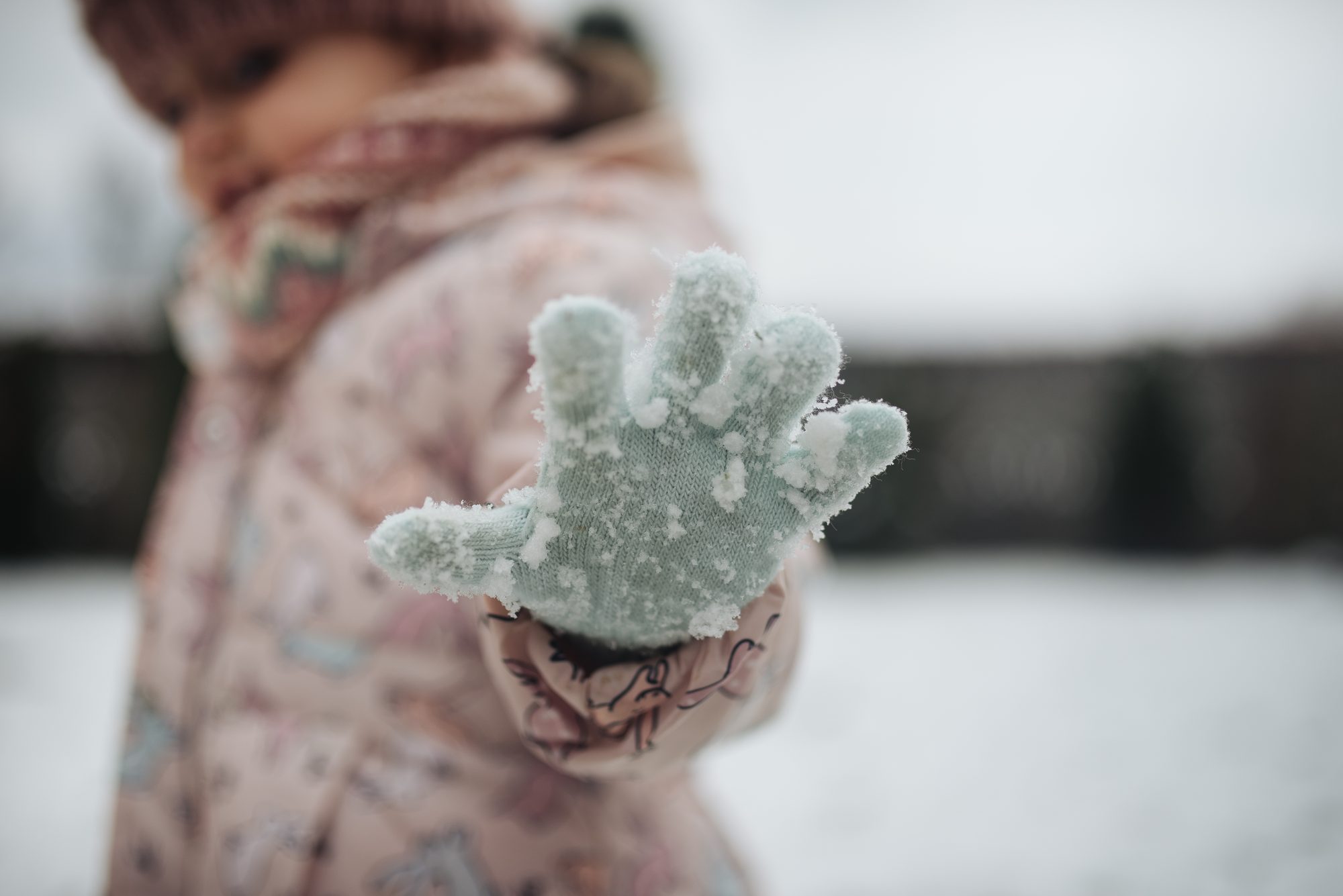 A young child in a winter coat and hat extends a gloved hand covered in snow while standing in a snowy outdoor area. The child radiates joy during a cold day of play.