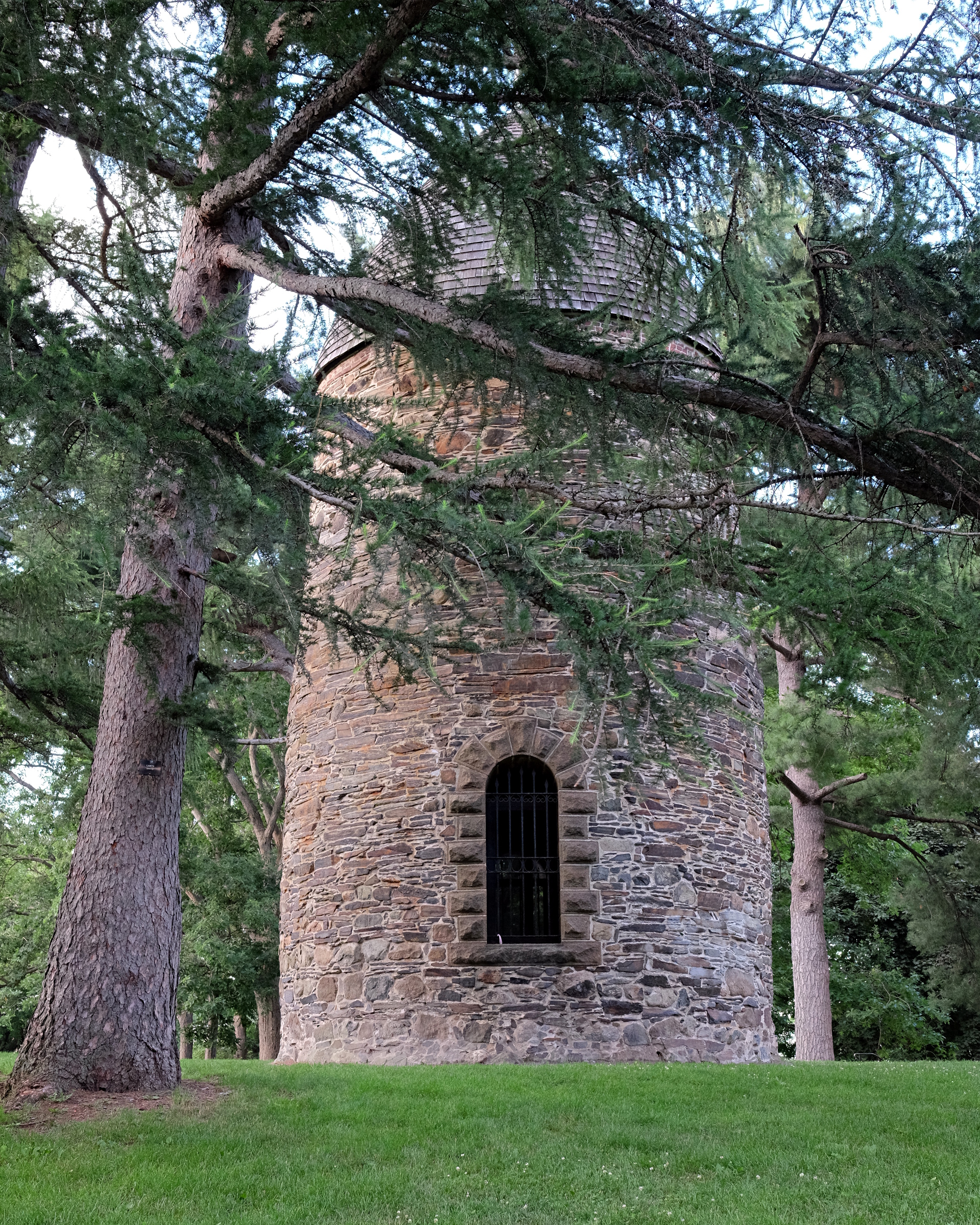 Cylindrical stone powder house tower with an arched barred window, framed by tall conifer trees.