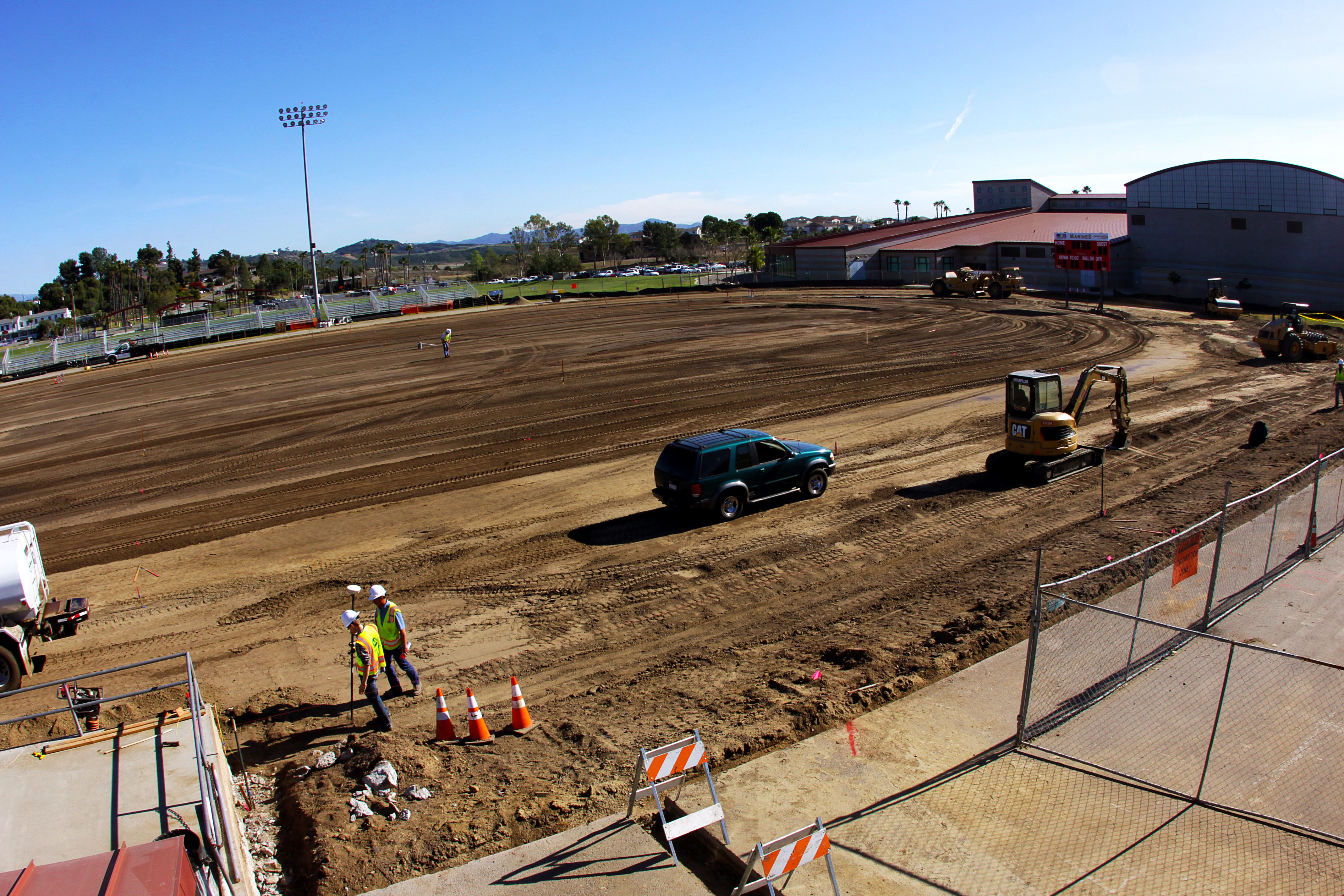 Dirt athletic field under construction with excavator, workers in safety vests, and chain-link fencing.