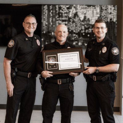 Three officers in black police uniforms pose together, center officer holding a framed certificate.