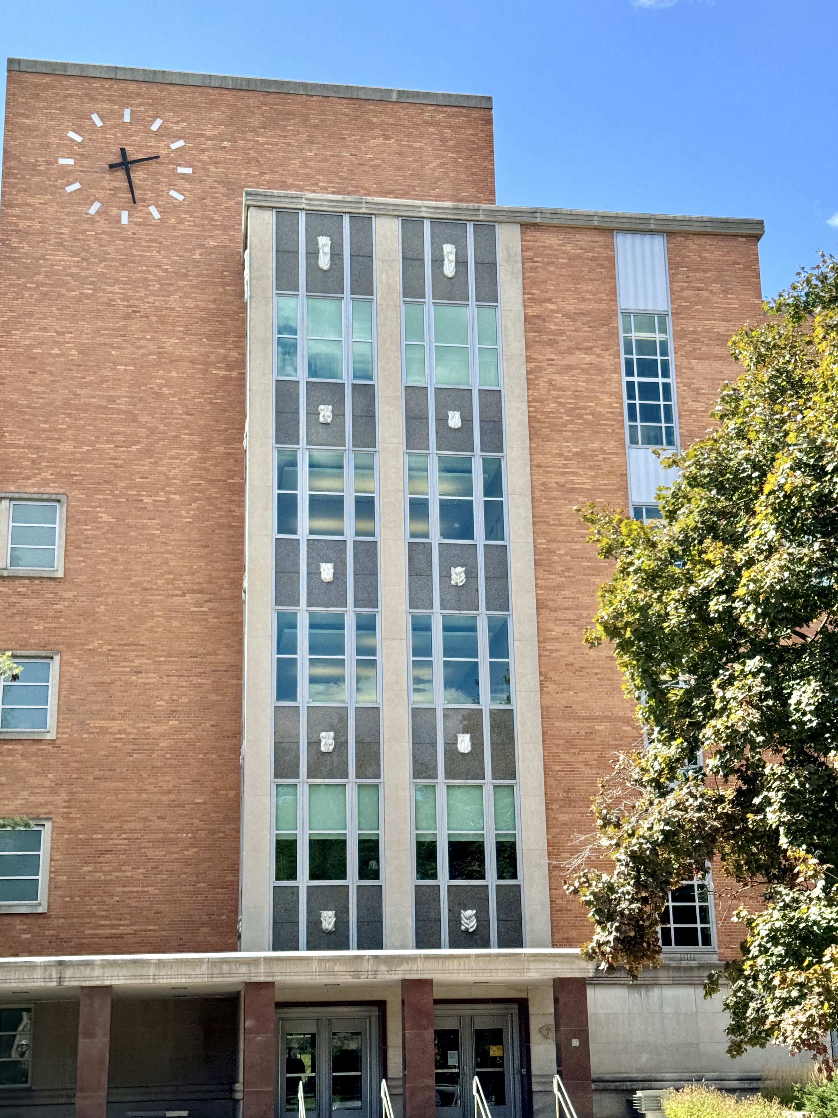 Mid-century brick university building with a large clock on its facade and tall vertical window bays.