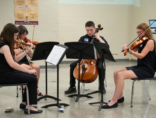 Four students in black attire play string instruments in a semicircle with music stands.