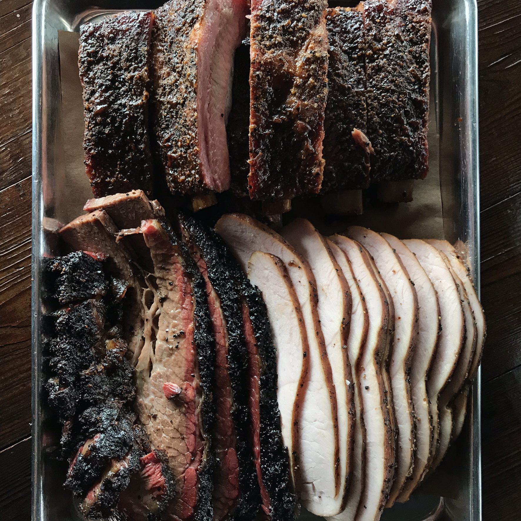 Metal tray of sliced smoked brisket, turkey, and beef ribs on a dark wooden surface.