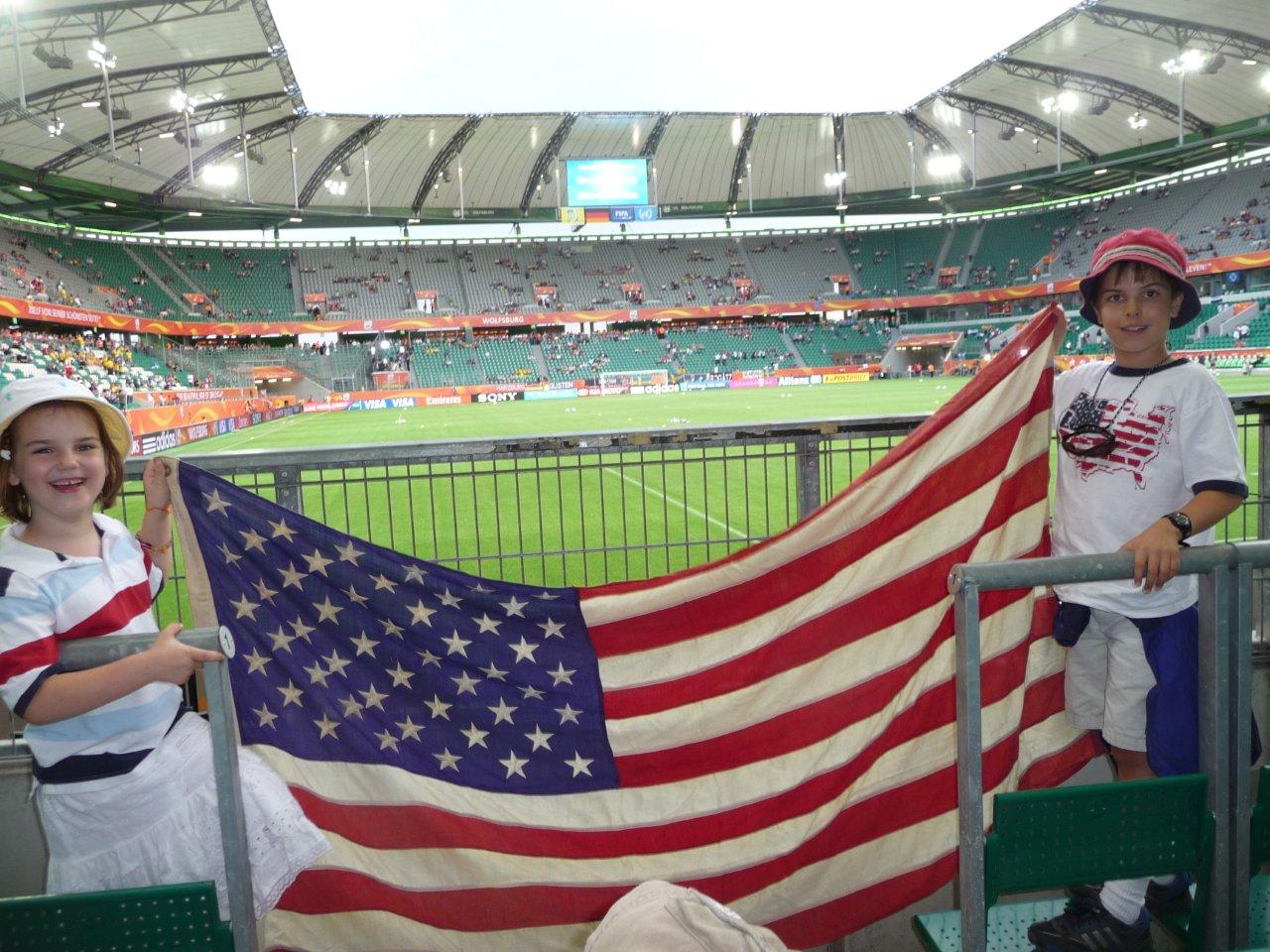 Two children hold a large American flag in a soccer stadium's stands.