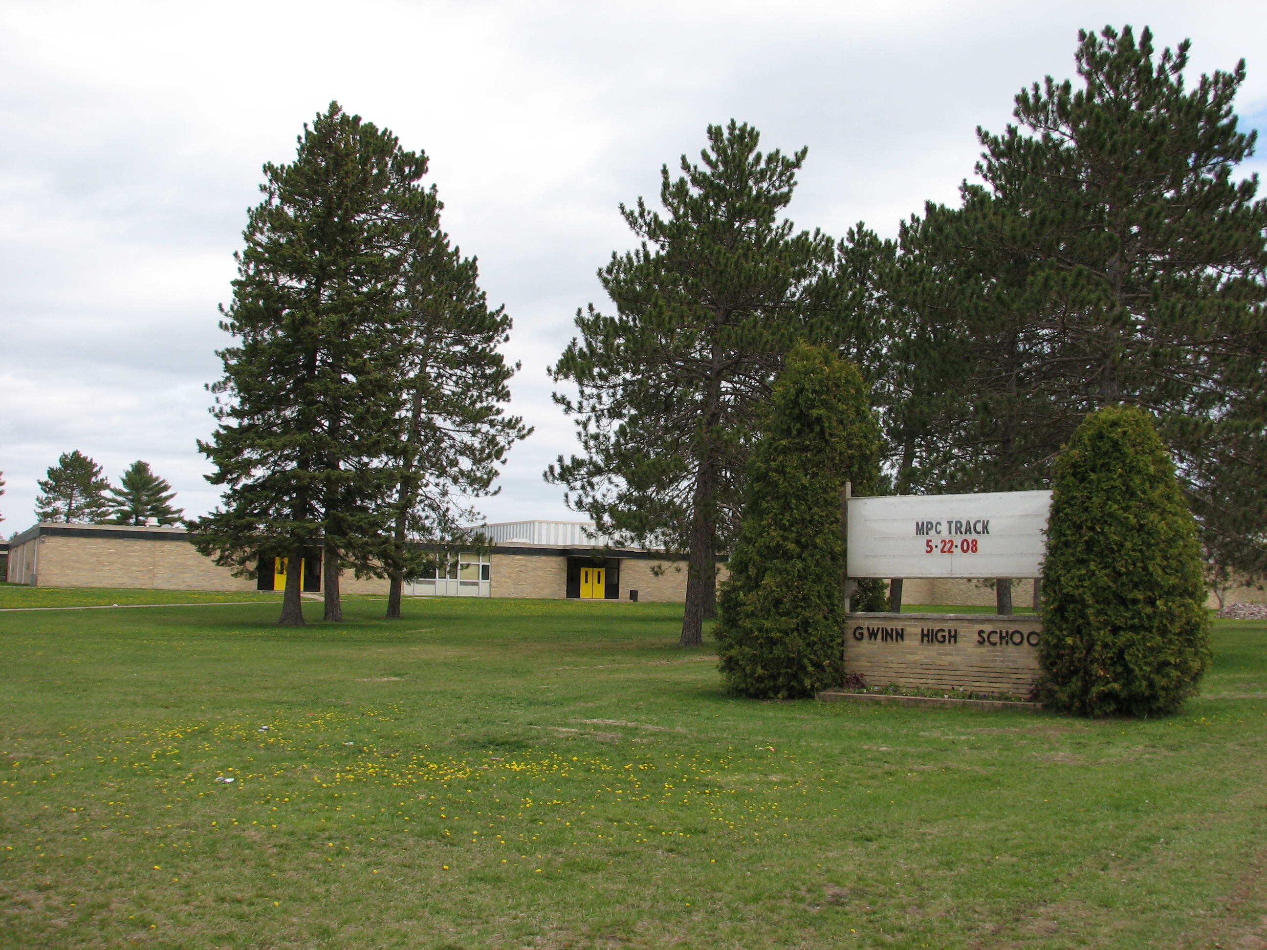 Single-story brick school building with a marquee sign reading Gwinn High School, surrounded by pine trees.