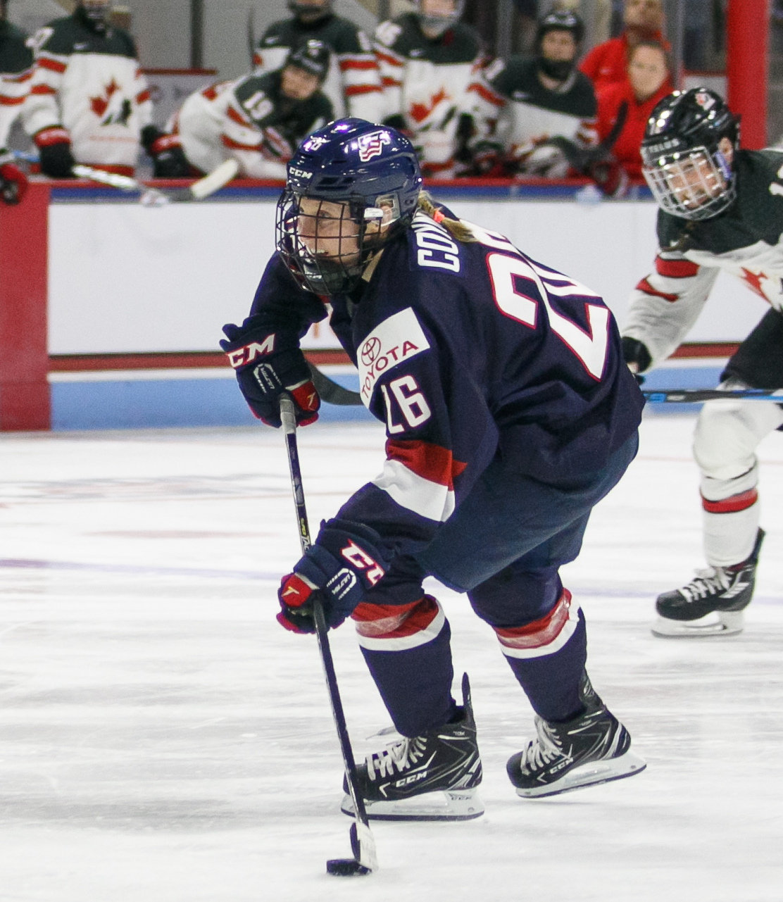 Female hockey player in a dark blue Team USA jersey number 26 skating with the puck on ice.