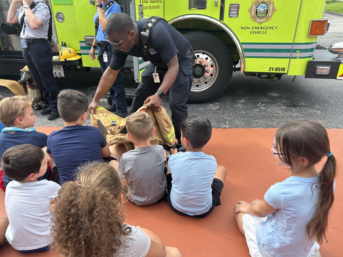 Firefighter in uniform shows gear to children seated in front of a yellow-green fire truck.