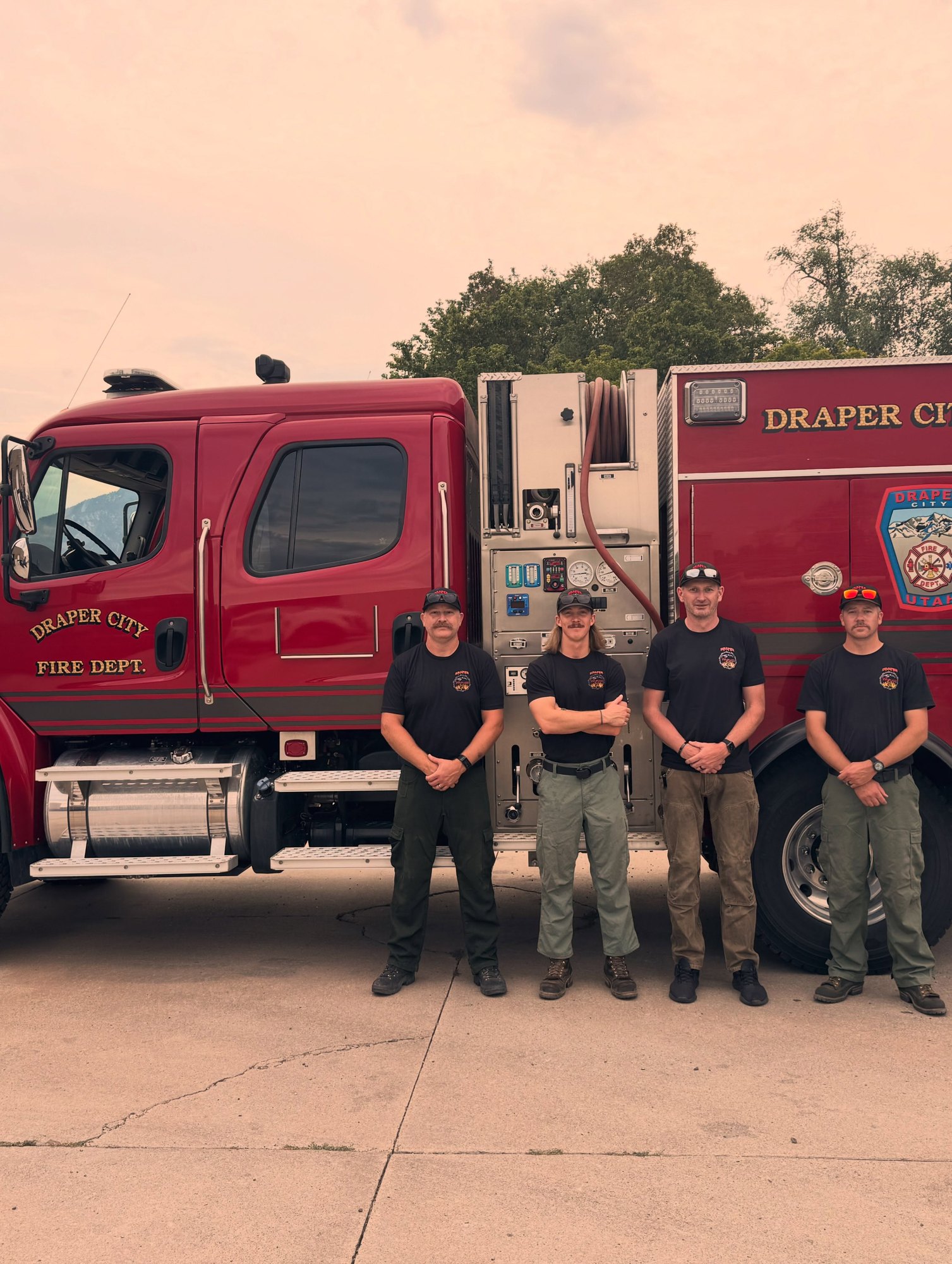 Four firefighters in dark shirts stand in front of a red fire engine marked Draper City Fire Dept.