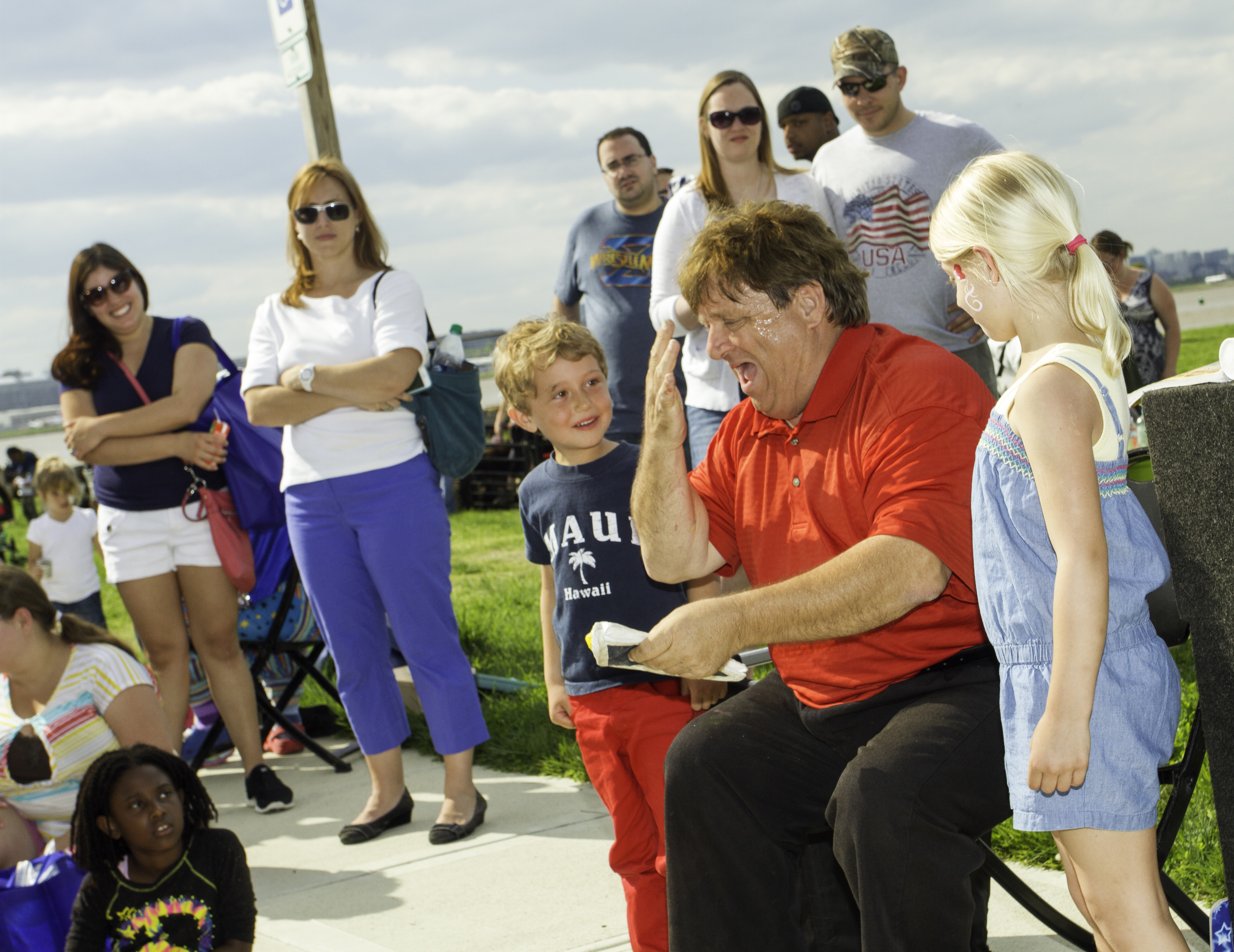 A performer in a red shirt crouches to interact with a child at an outdoor family show.