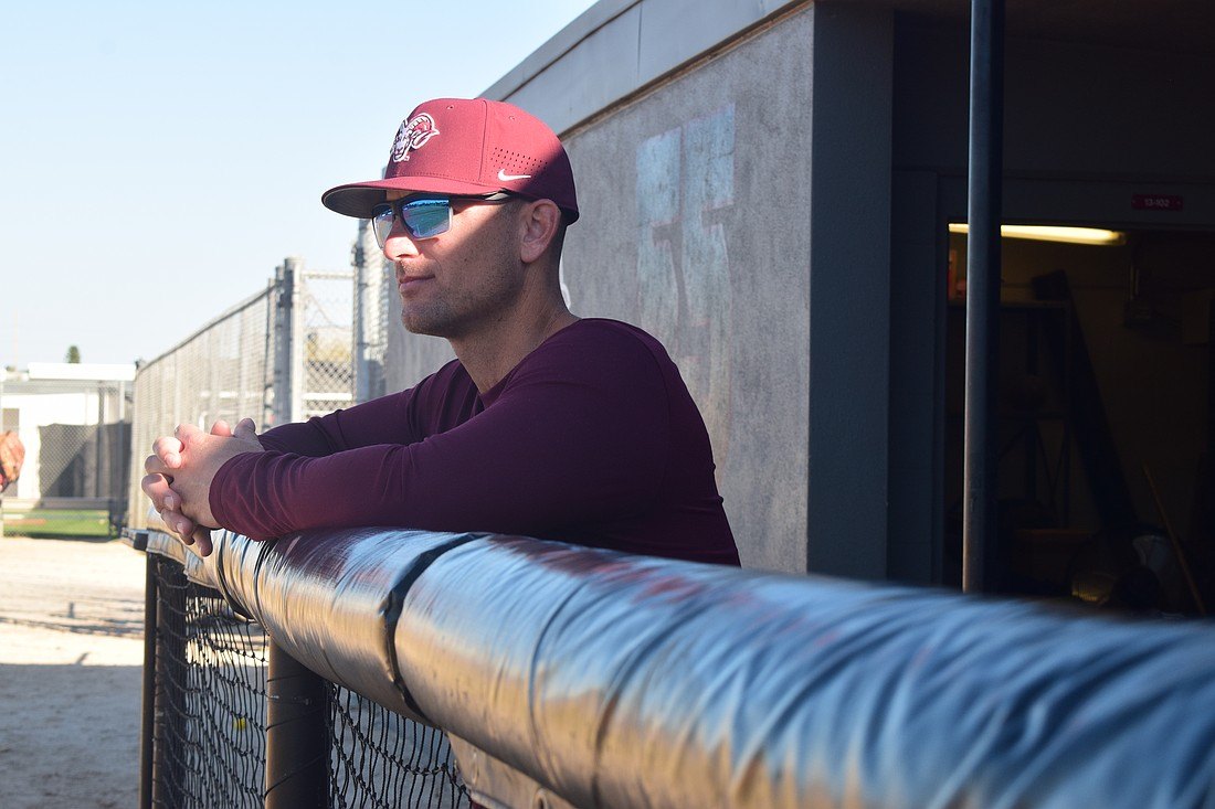 A man in a maroon shirt and red baseball cap leans on a dugout railing at a baseball field.