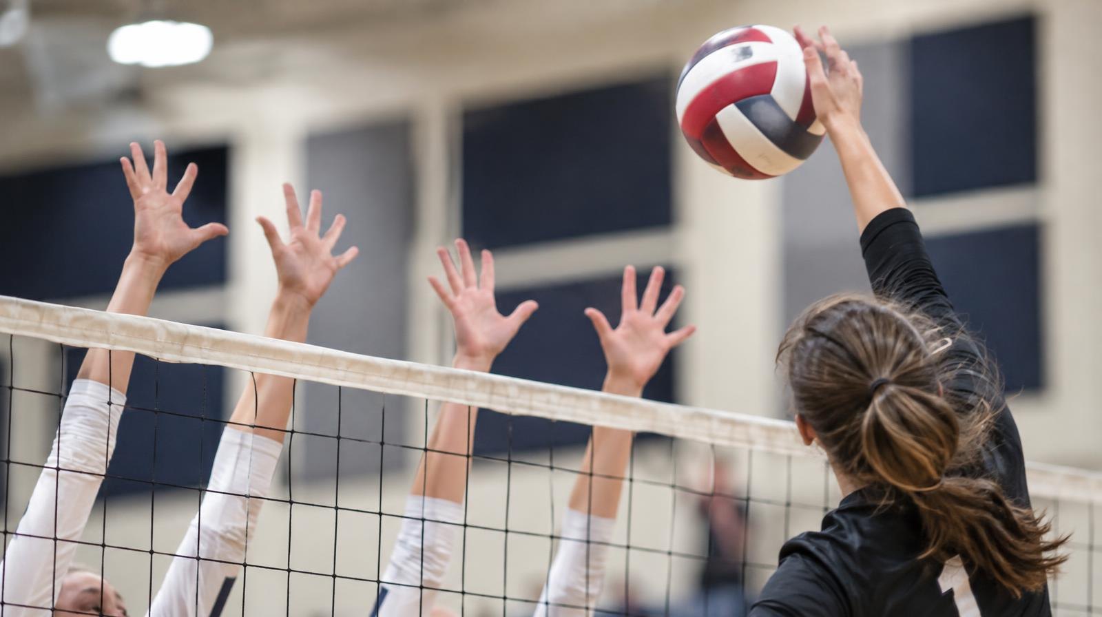 A volleyball being spiked over a net with players' hands reaching up to block, indoors.