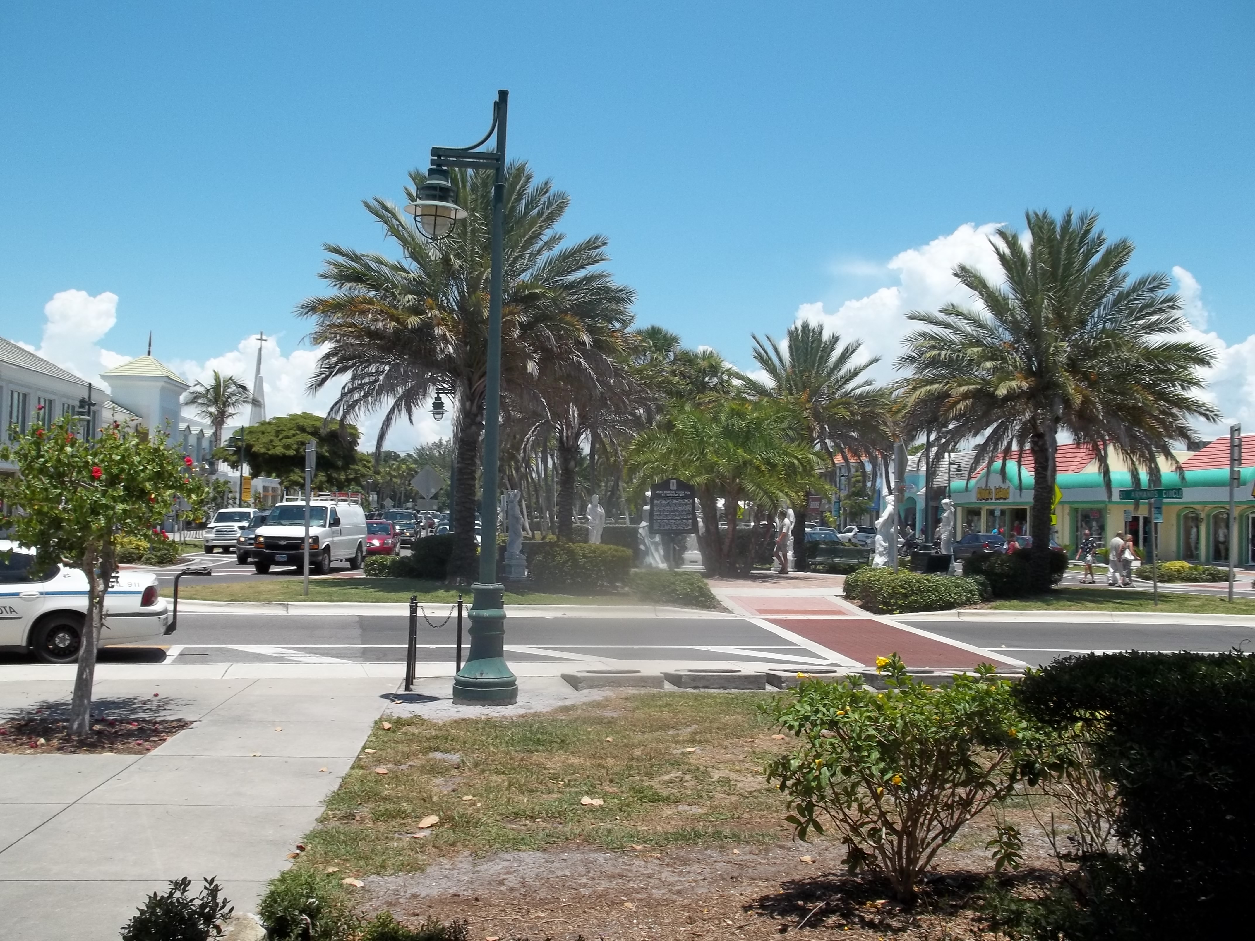 Palm trees and a green ornate street lamp line a sunny commercial boulevard with storefronts.