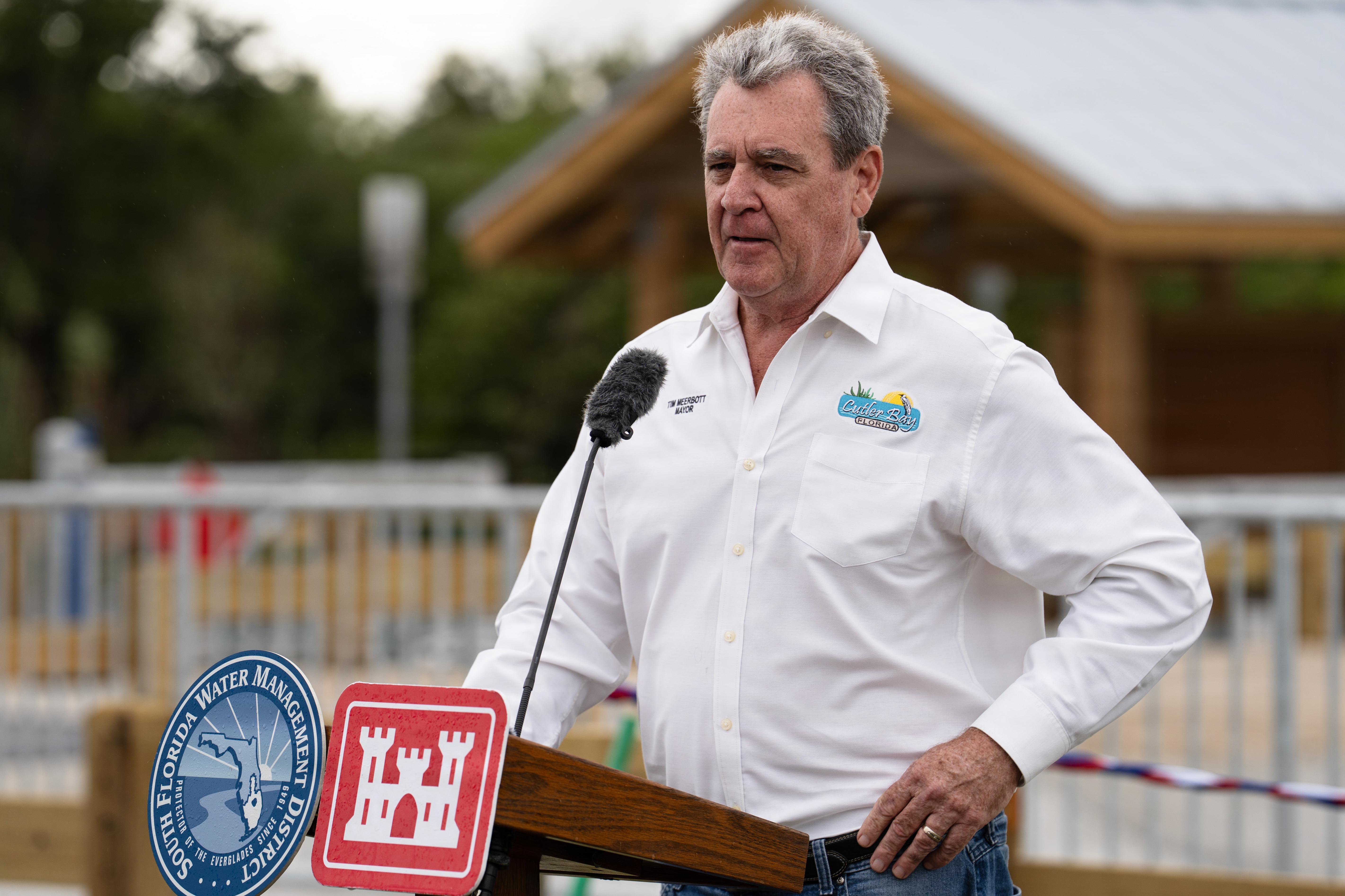 Man in white shirt speaks at a podium with agency seals displayed at an outdoor event.