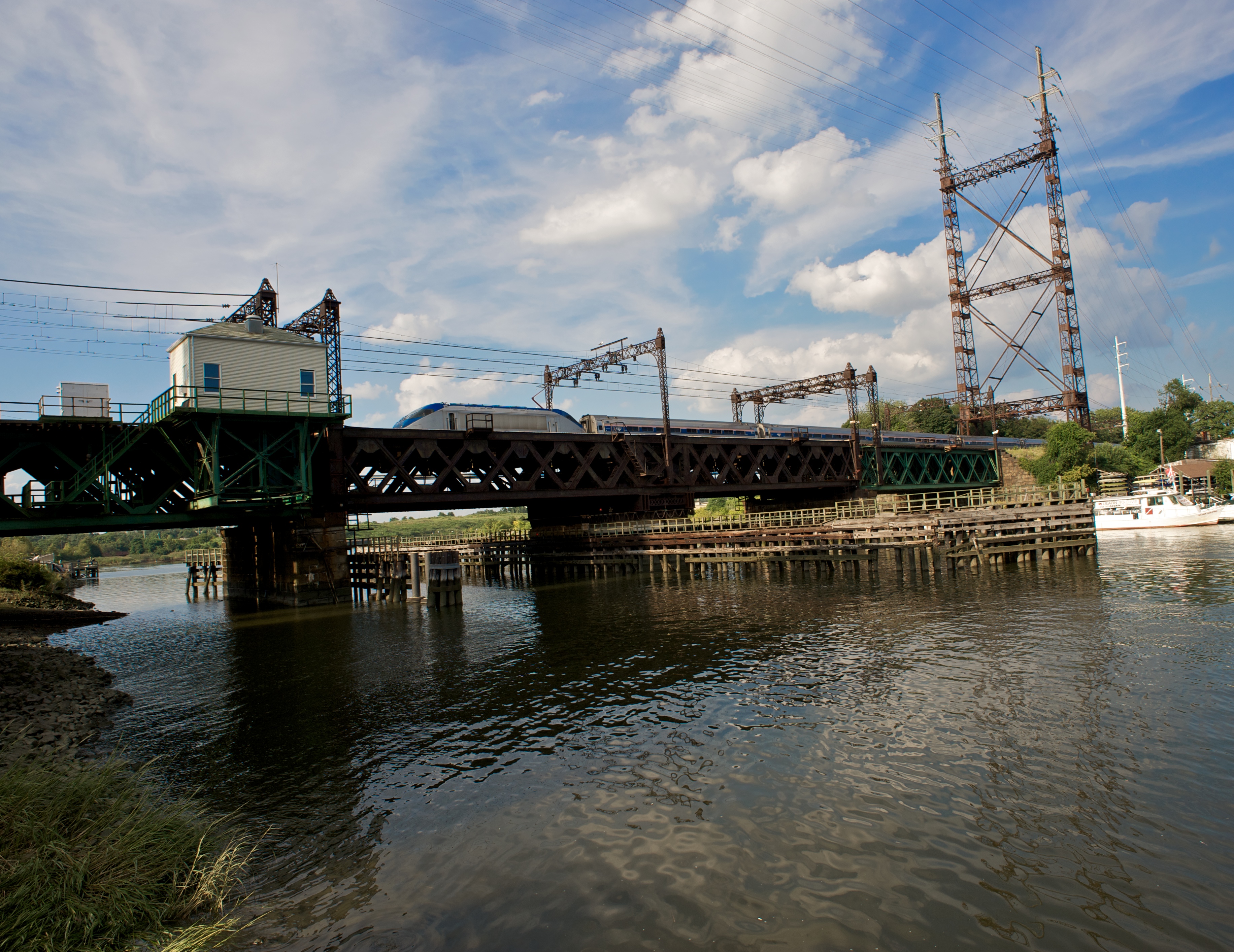 A passenger train crossing a green steel railroad bridge over a river with catenary wires overhead