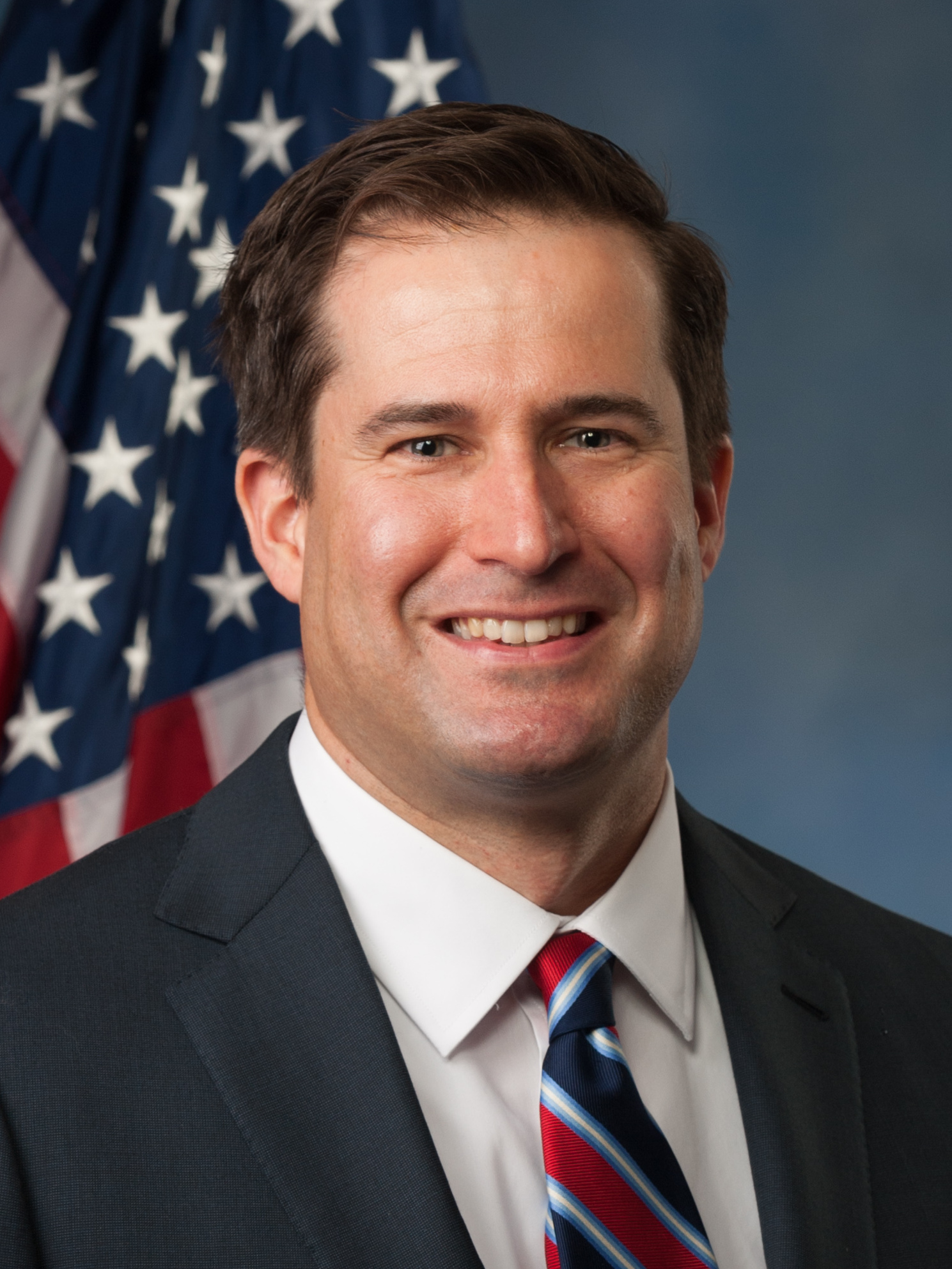 Man in dark suit and striped tie smiling before an American flag.