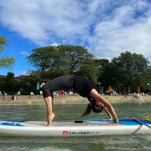 Woman doing a wheel pose on a paddleboard on calm water with trees and beach umbrellas in background.