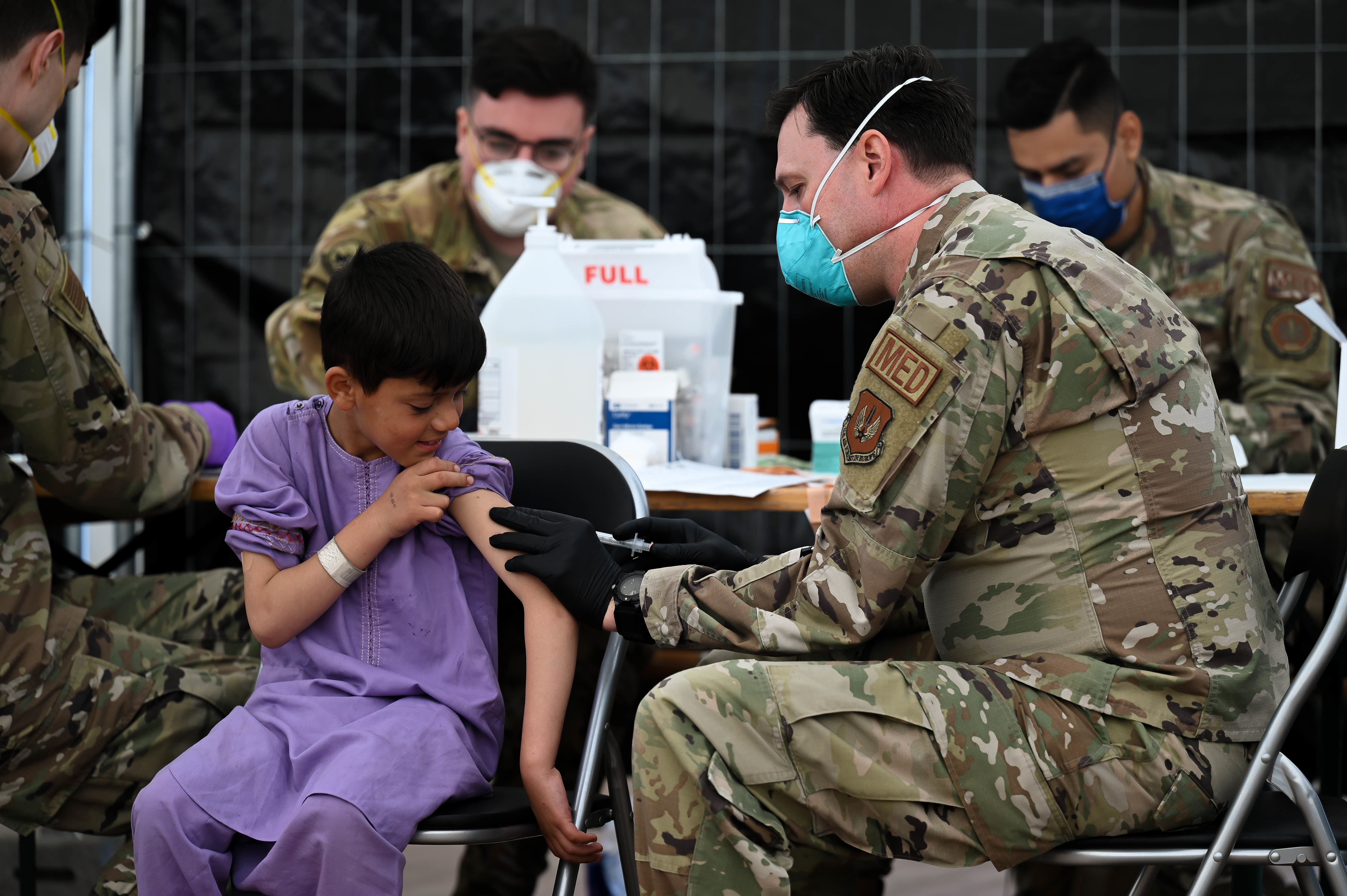A uniformed airman administers a vaccine to a young child at an indoor vaccination station.