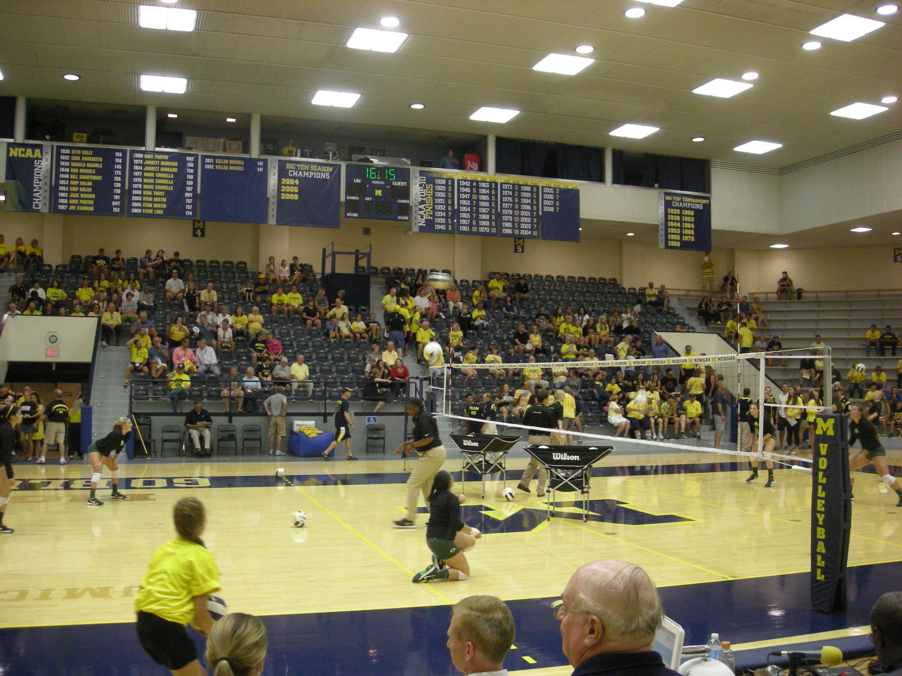 Players compete on a volleyball court inside an arena with a maize-and-blue crowd.