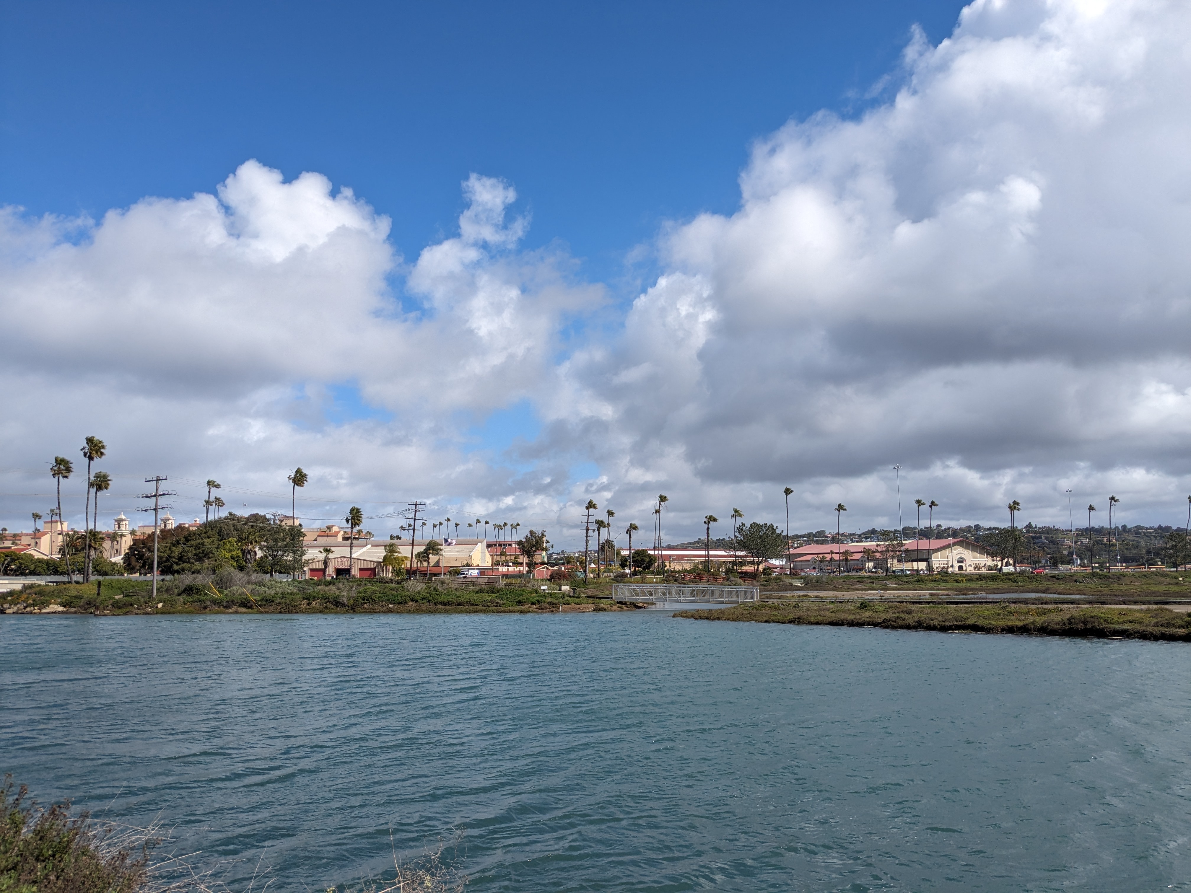 Fairgrounds buildings and palm trees reflected across a calm lagoon under blue sky.