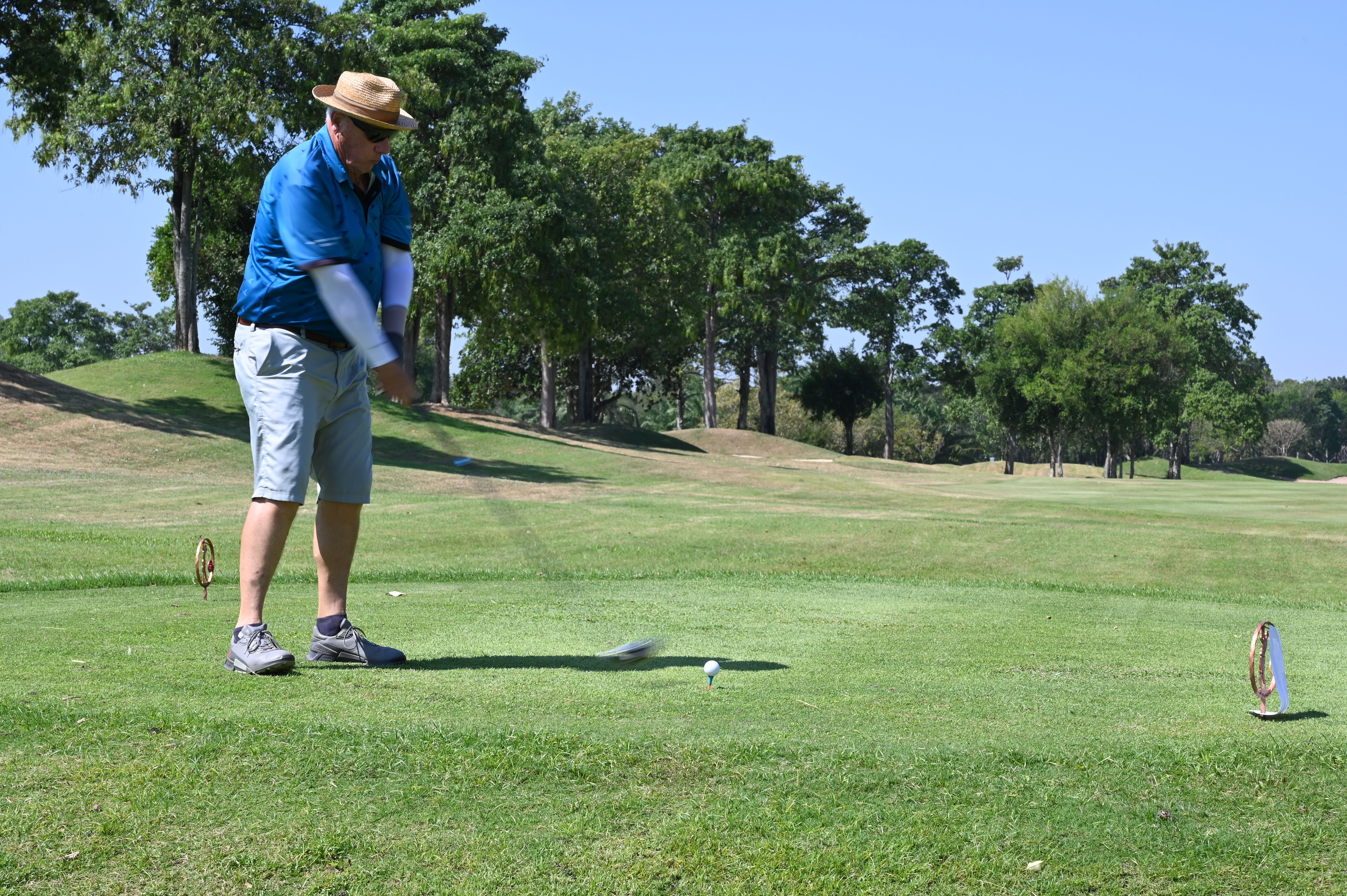 A golfer in a straw hat swings a driver mid-tee-shot on a sunny fairway.