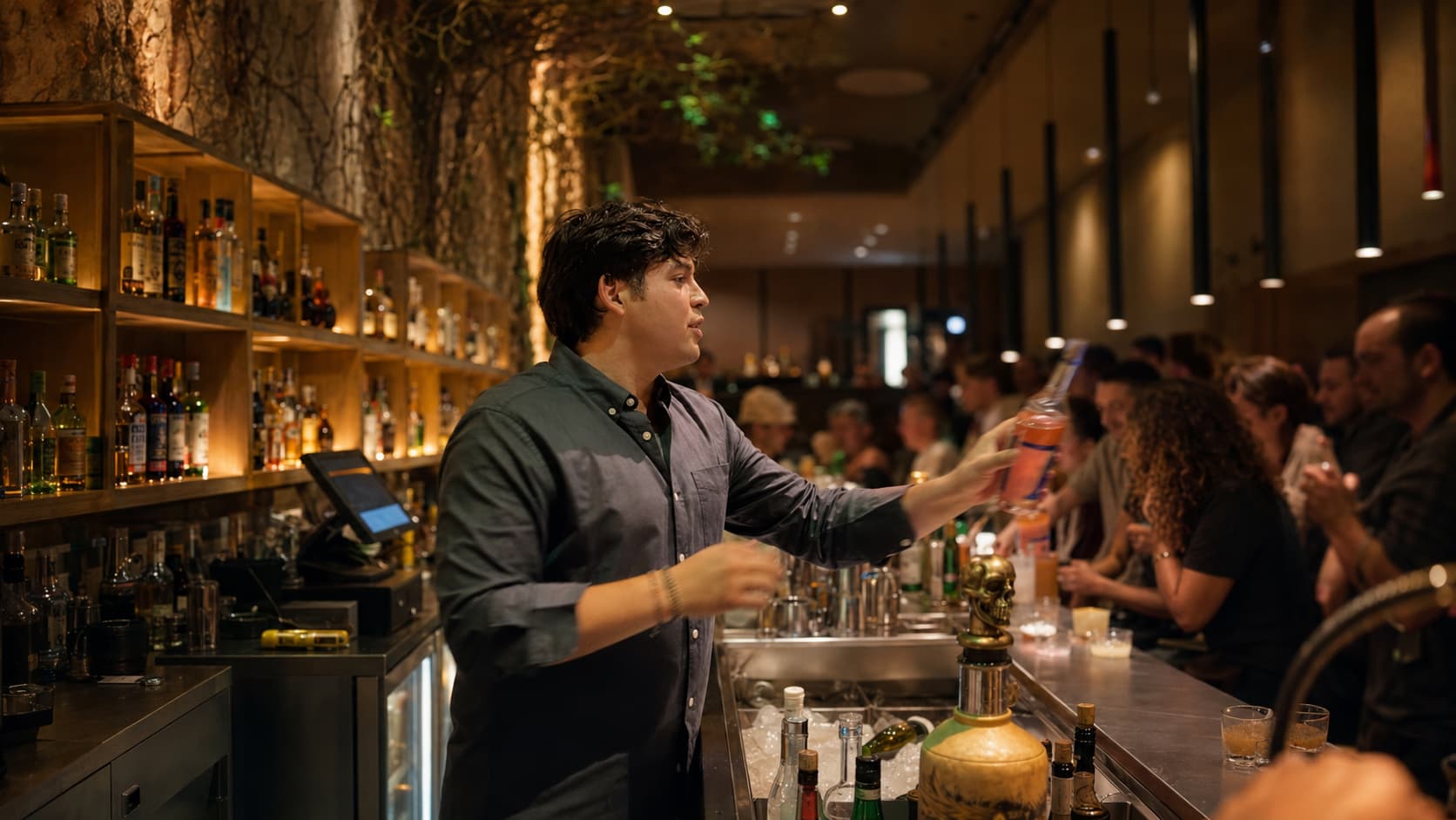 A bartender reaches across a crowded bar stocked with backlit spirits bottles.