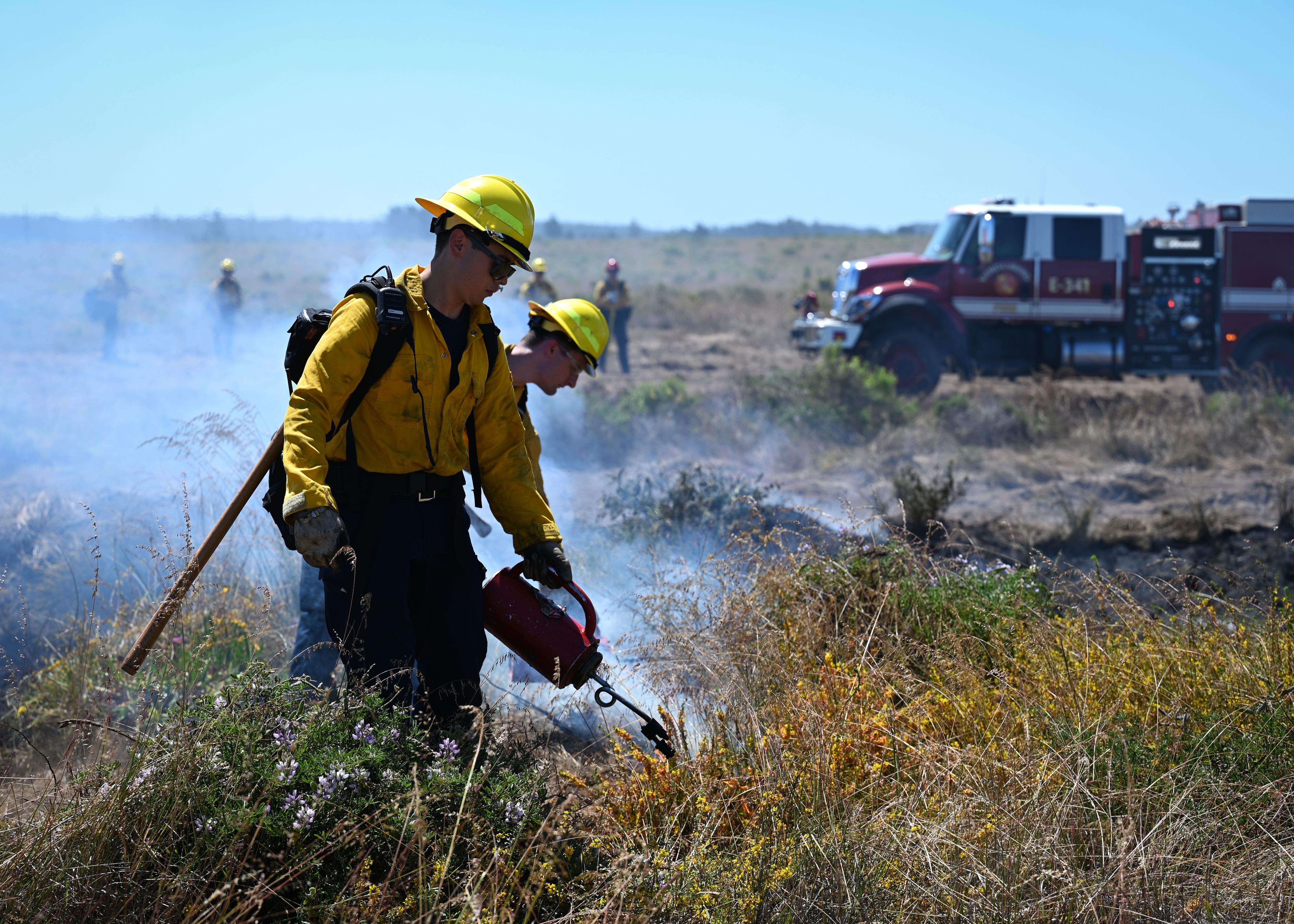 Firefighter in yellow gear igniting dry scrub brush with a drip torch, smoke rising nearby.