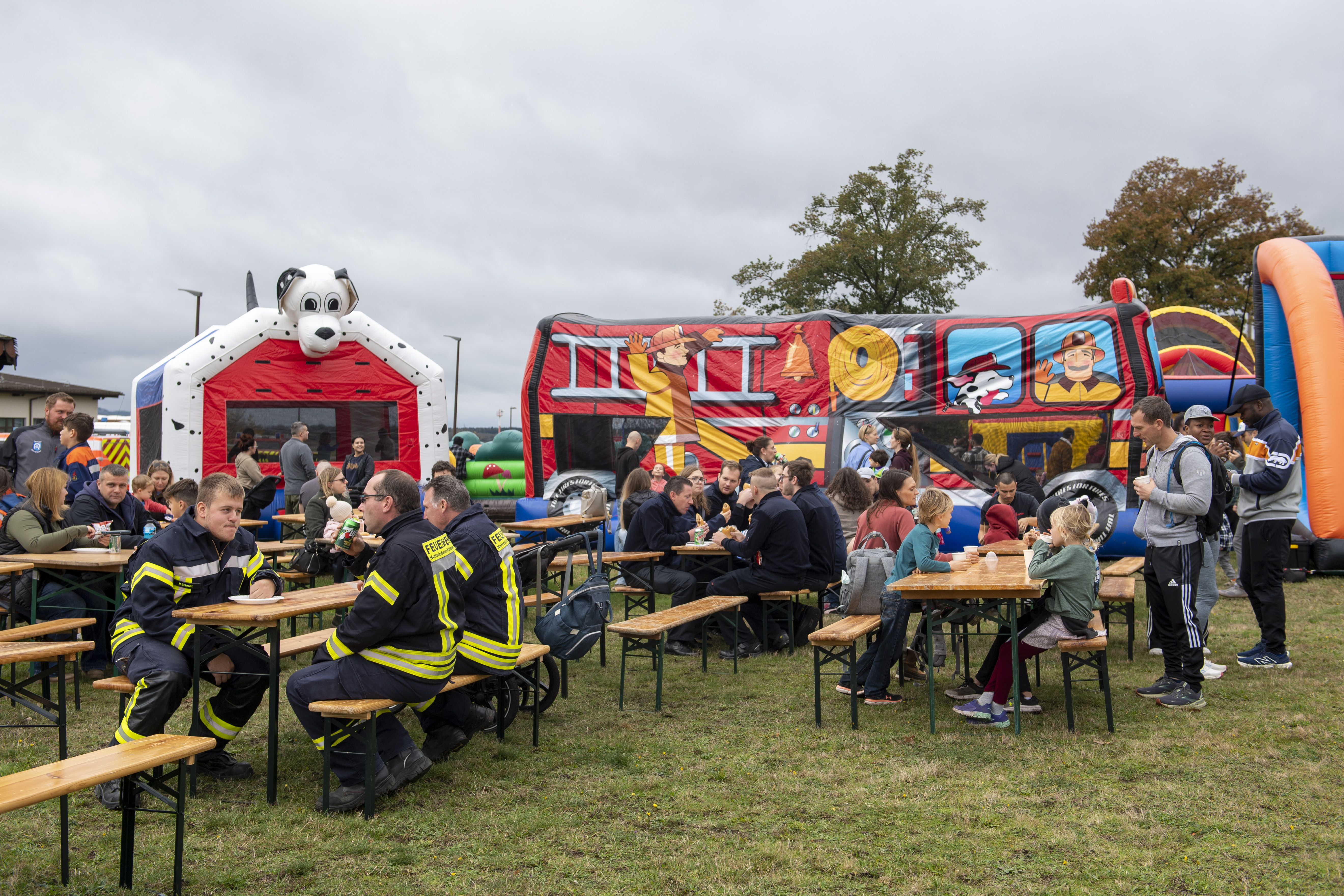 Firefighters and families seated at picnic tables at an outdoor fire station open house.