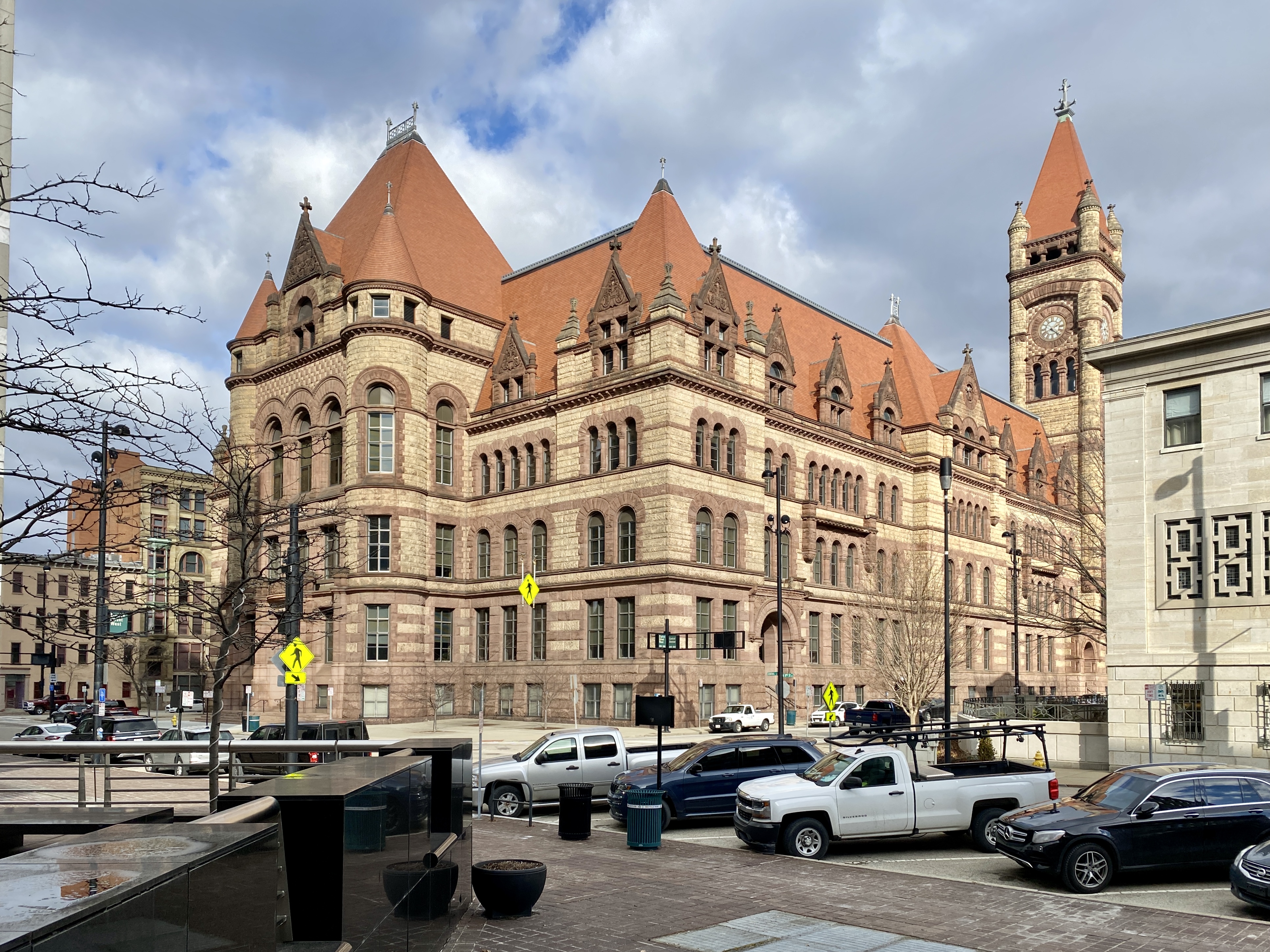 Wide view of a Romanesque stone city hall building with a clock tower and parked vehicles.