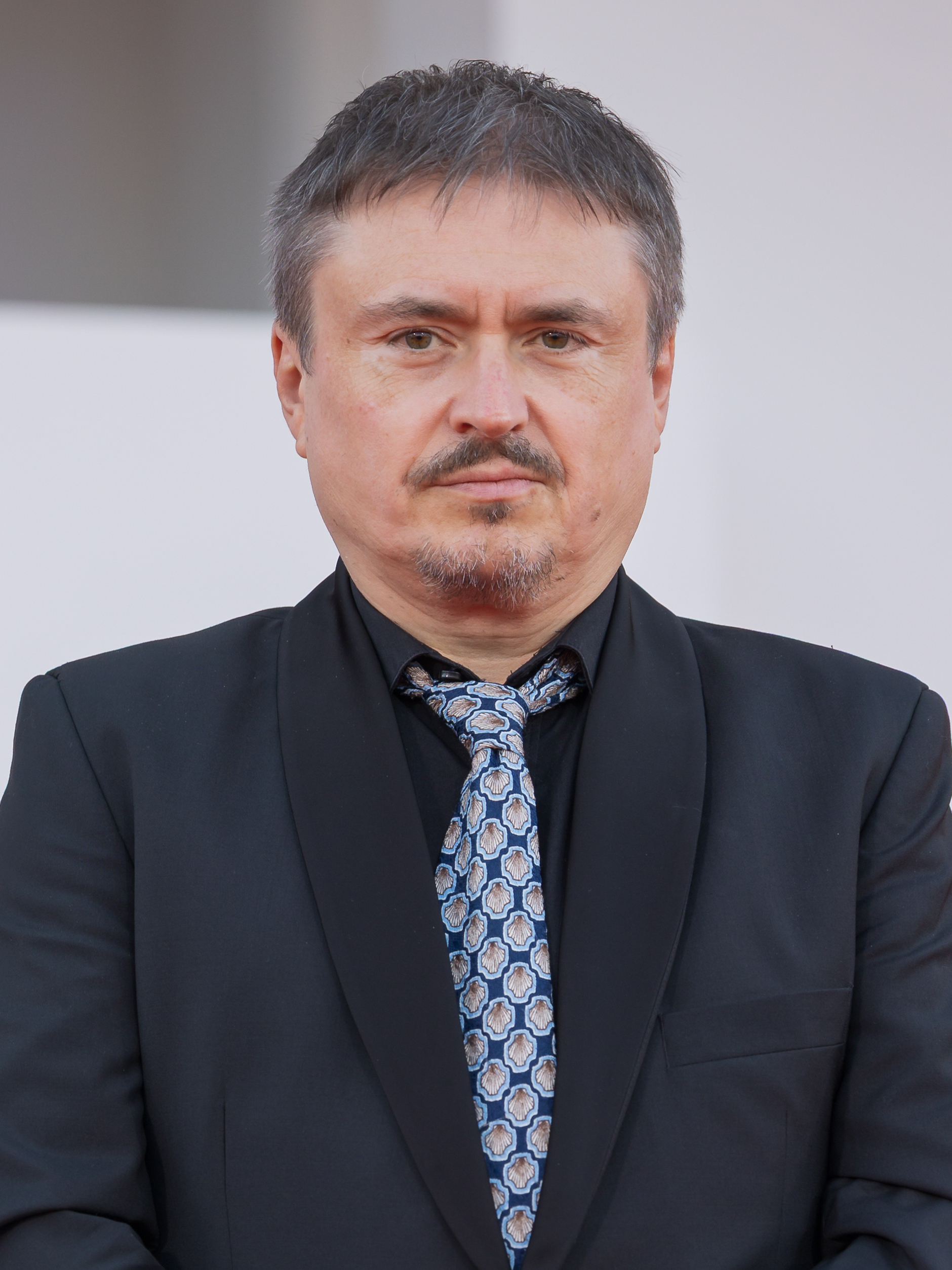 Man in black suit and patterned tie standing against a light backdrop.