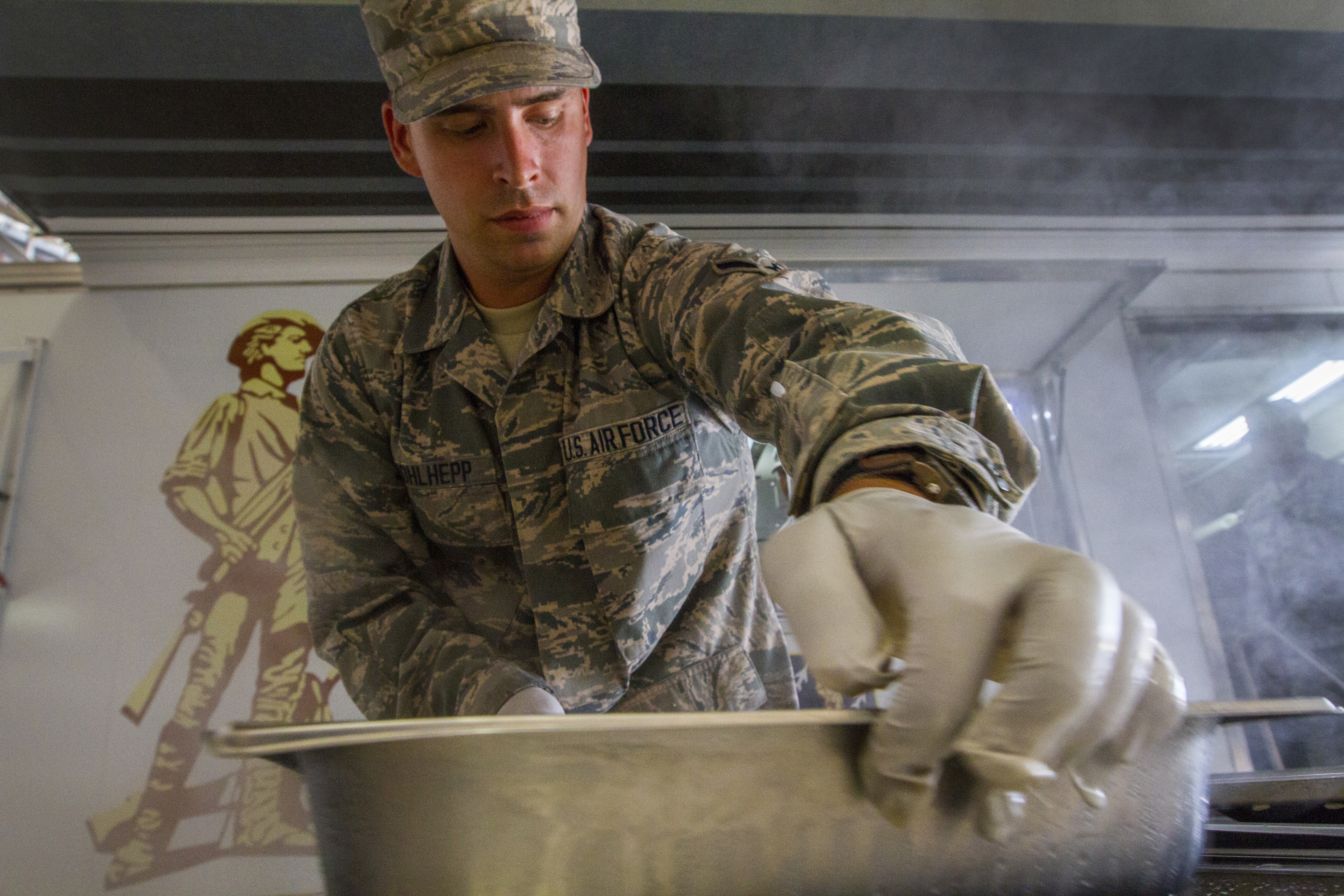 A uniformed airman in gloves stirs food inside a mobile kitchen trailer with steam rising.