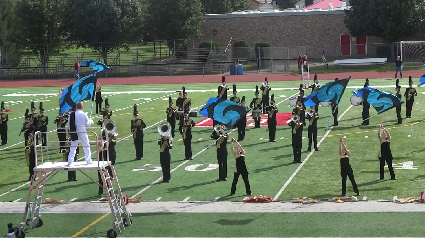 Marching band performs on a football field with color guard waving large teal flags.